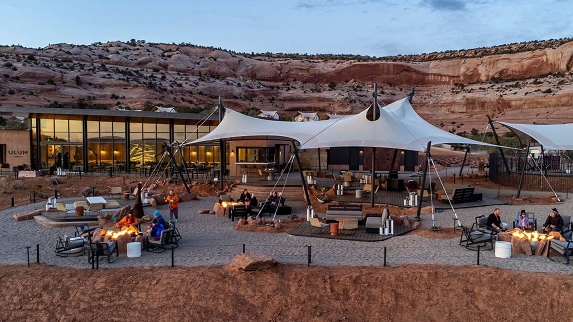 Desert resort with open-air dining under large tents, fire pits, and a modern building at dusk.