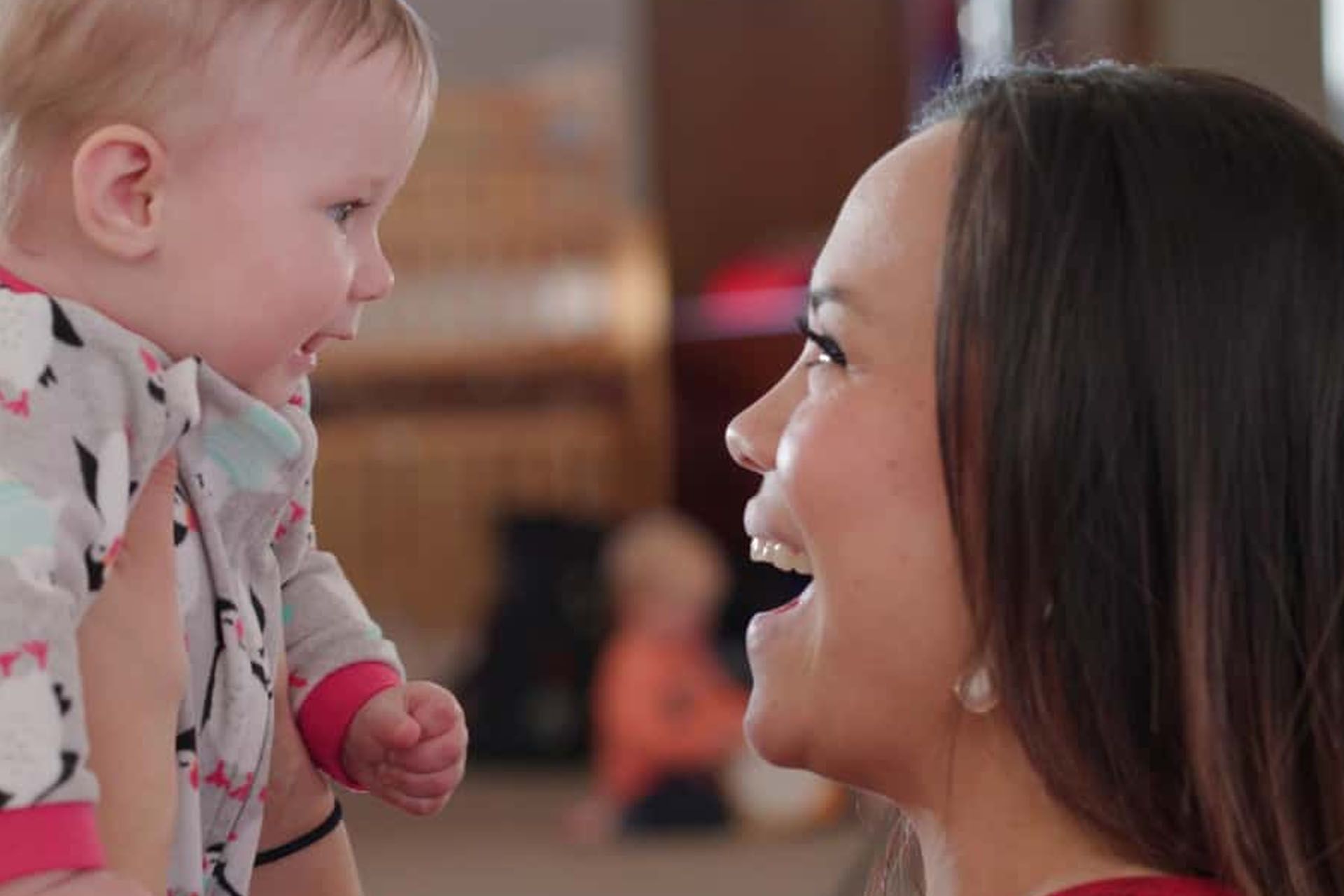 Woman holding baby, both smiling face-to-face. Indoors with blurred background.
