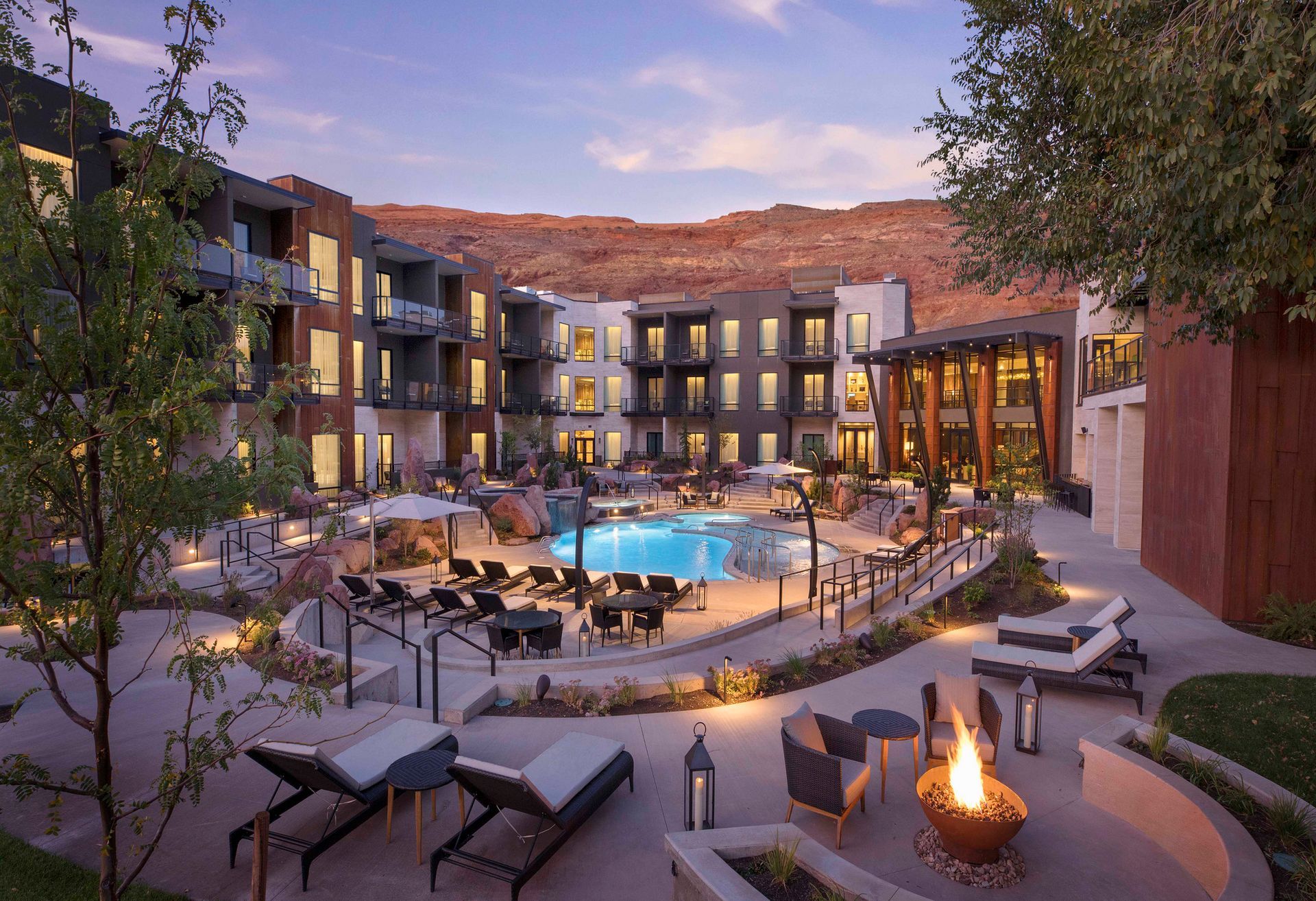 Resort courtyard with pool, lounge chairs, fire pit, and red rock mountains in the background.