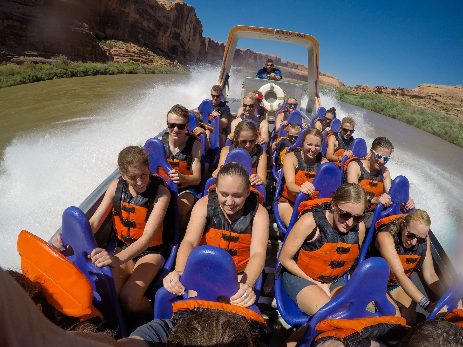 People on a speeding river boat, water splashing, against a desert canyon backdrop.