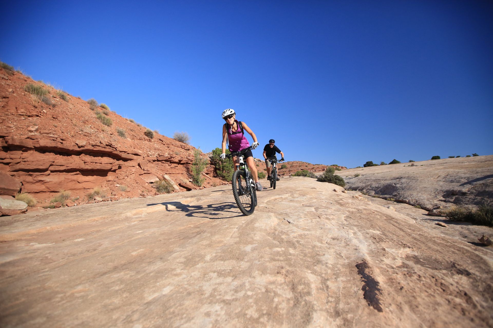 Two people mountain biking on a sandy trail under a bright blue sky. Red rock cliffs in the background.