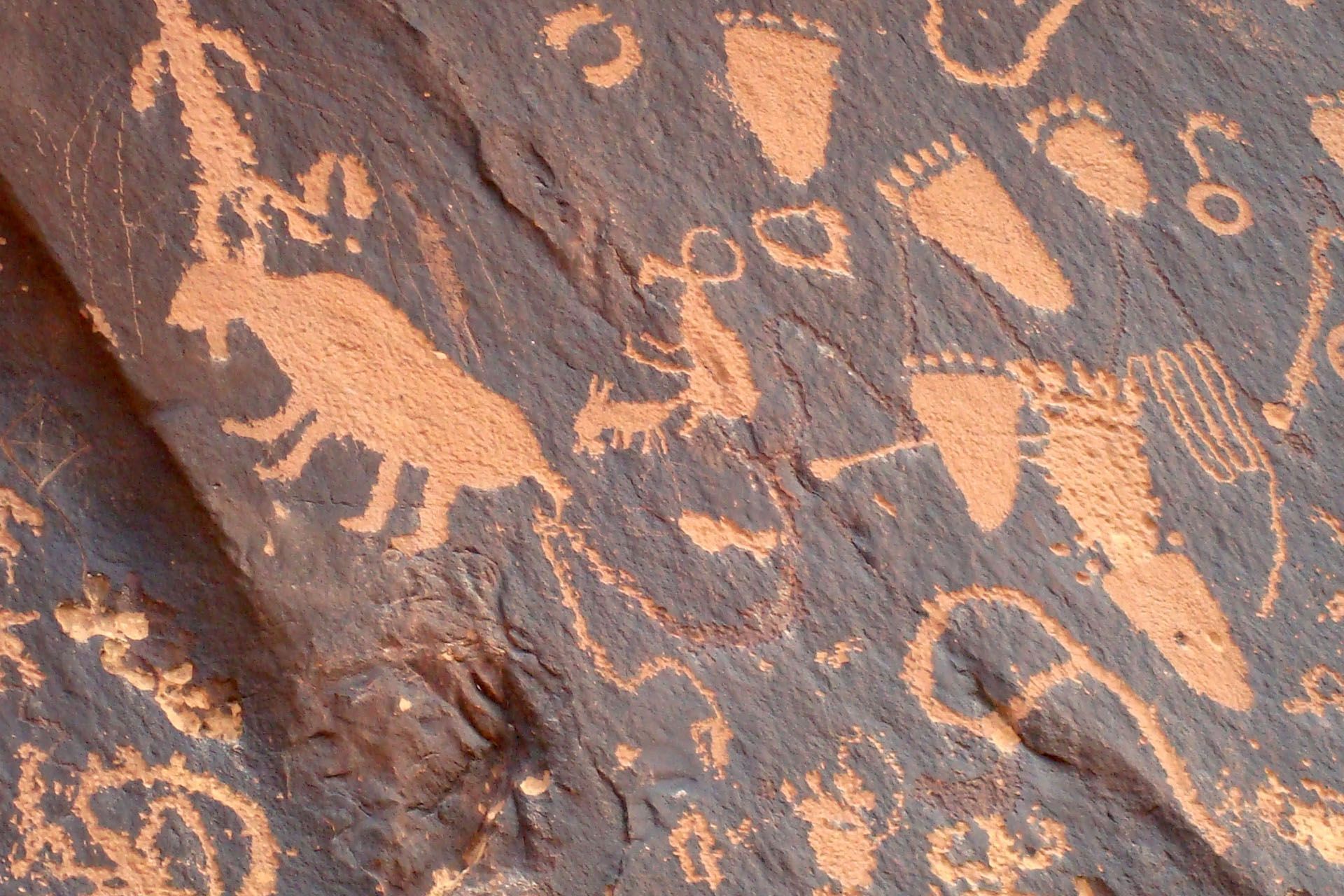 Rock art with reddish-brown petroglyphs of figures, animals, and symbols on a dark rock surface.