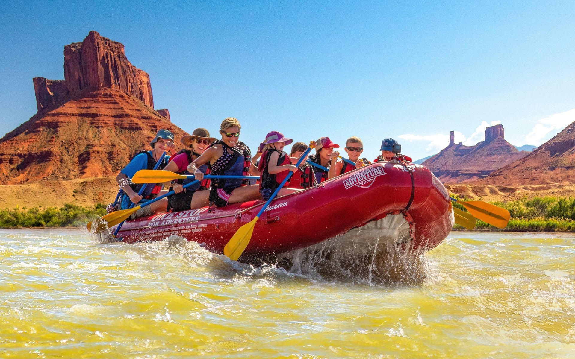 Rafting on a river with a group of people, red raft with paddles, red rock formations in background.