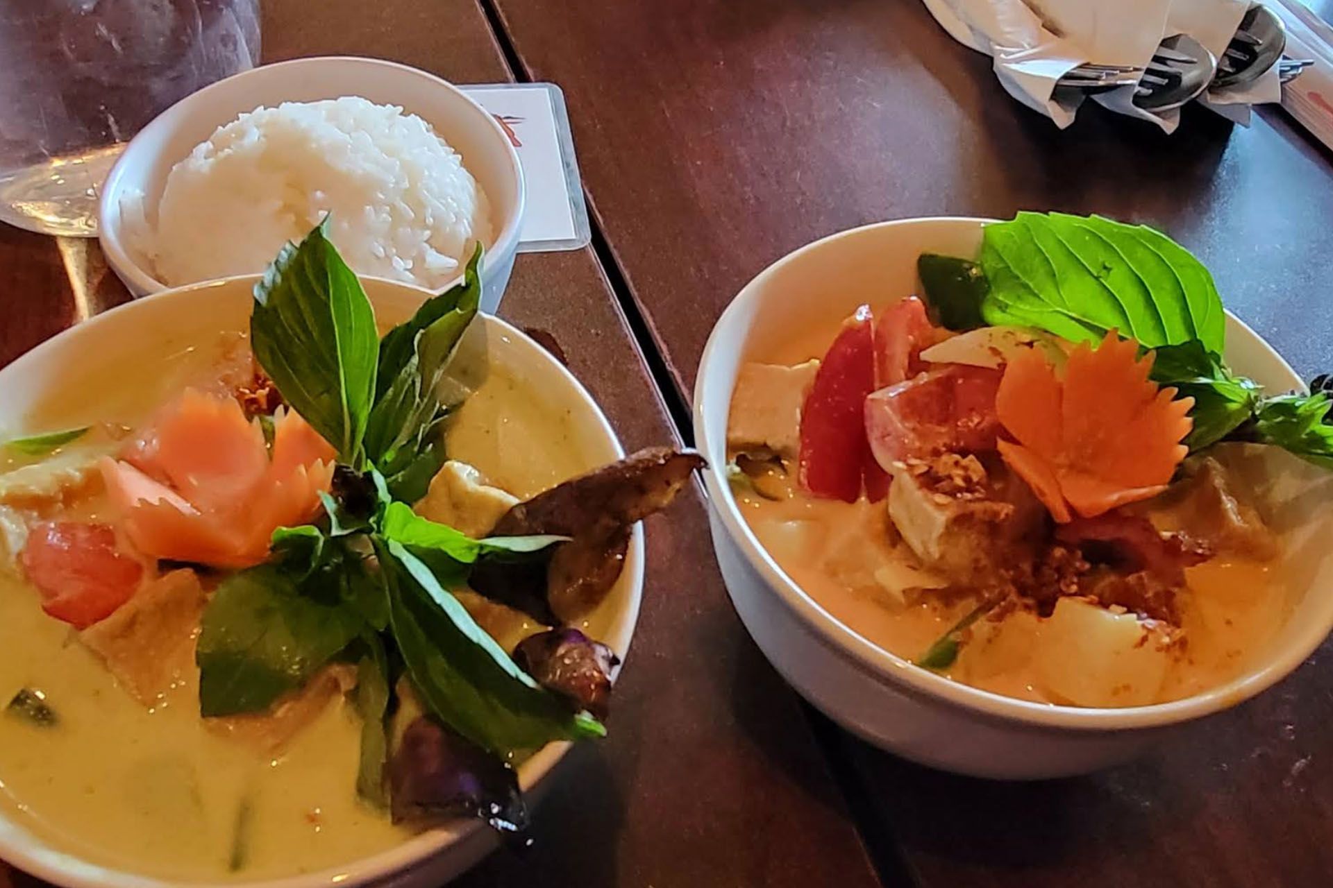 Two bowls of Thai curry, rice, and water on a wood table. One green, one red. Garnished with vegetables.