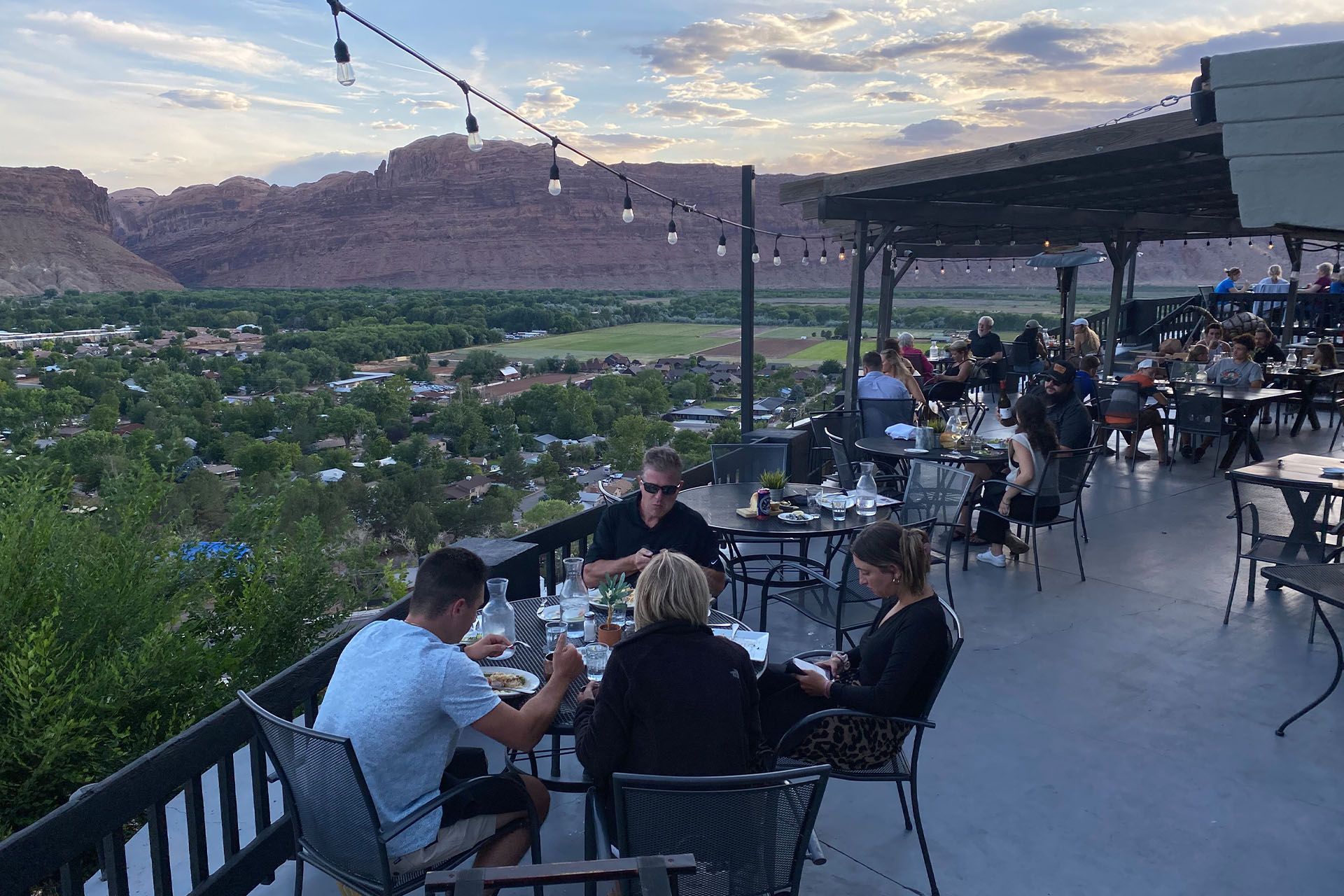 Restaurant patio with diners overlooking a town and red rock mountains at sunset.