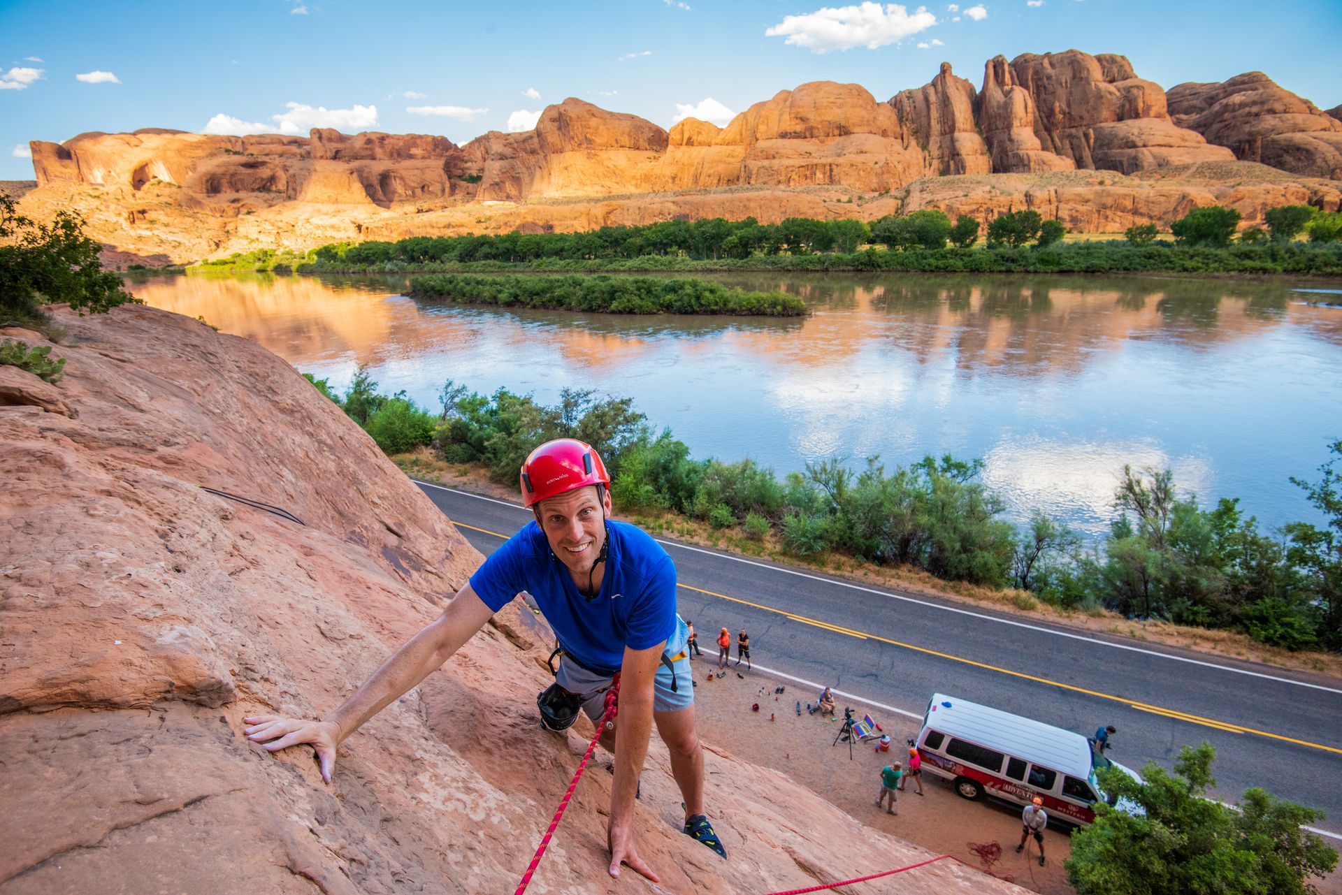 Rock climber in blue shirt and red helmet scales a sandstone cliff, river and road below.