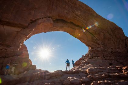 Two pepole stand under a massive stone arch with the sun shining through the middle.