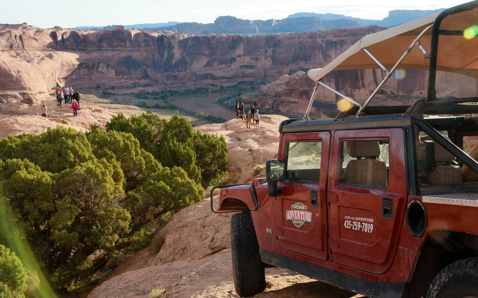 A family explores a dramatic overlook into the Colorado River with a hummer vehicle parked in the background.