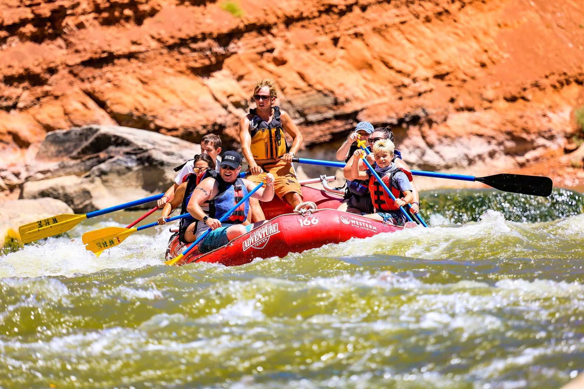Rafting on a river with several people paddling through rapids near red rock formations.