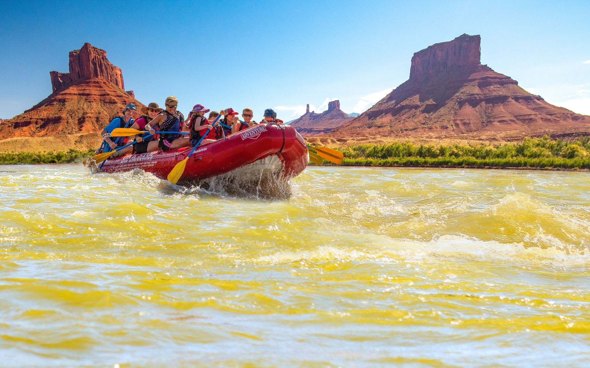 Rafting on a river with people, red raft, yellow water, red rock mountains in the background, blue sky.