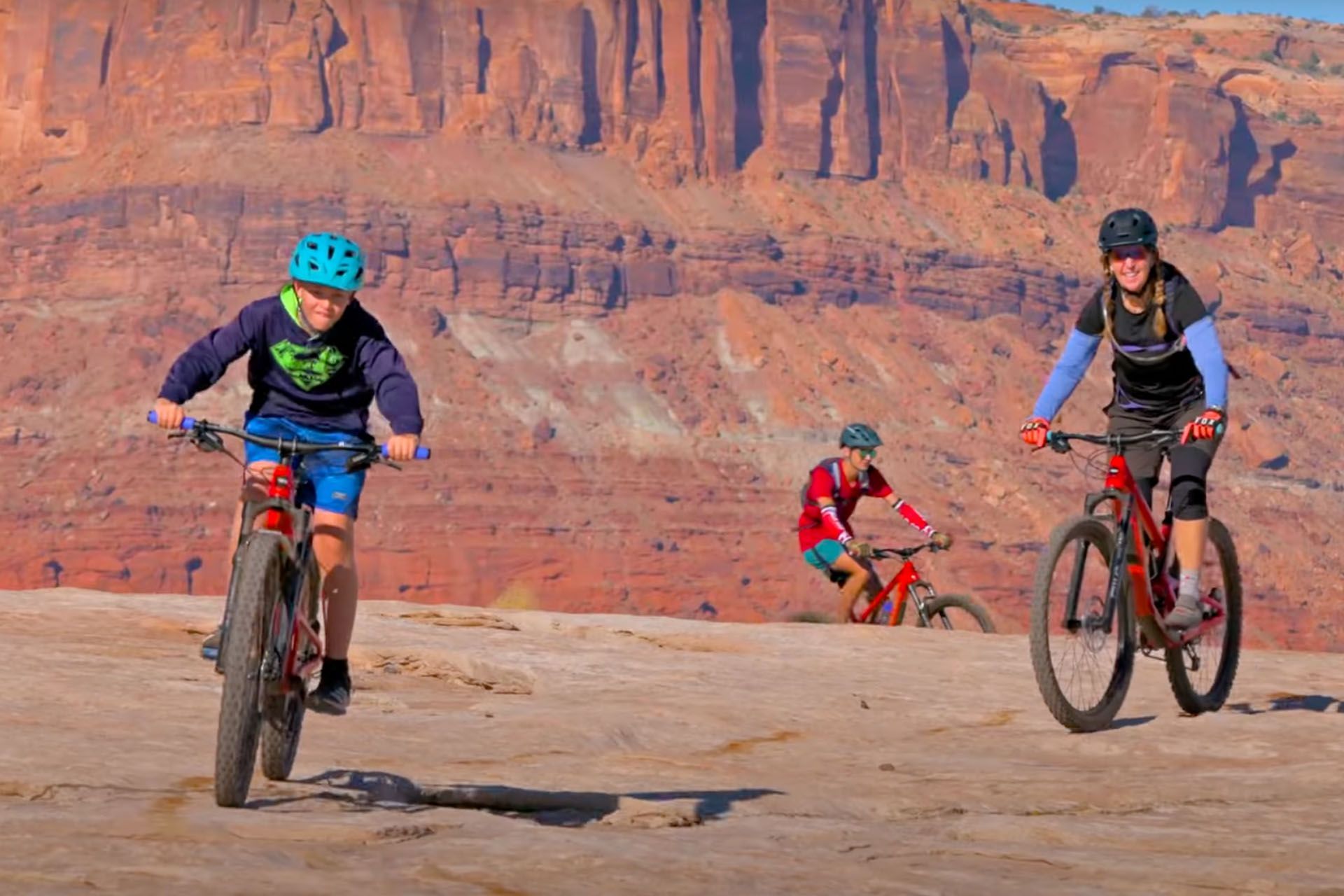 Three mountain bikers ride across a desert landscape with red rock cliffs.