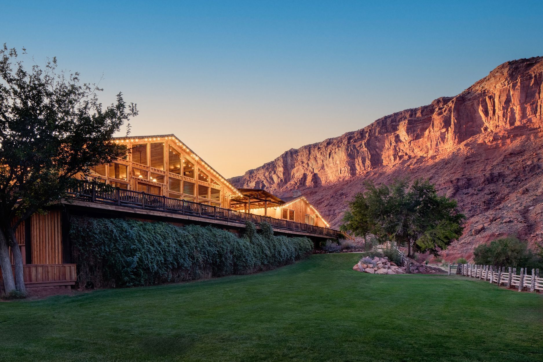 A resort building with wooden beams and a green lawn at the foot of a dramatic cliffs at sunset.