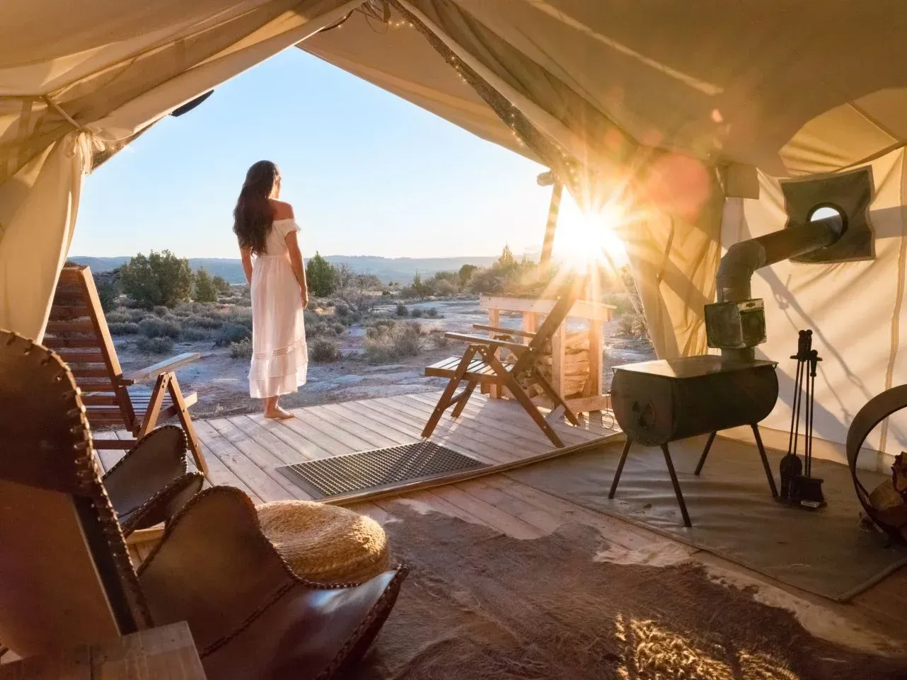 Woman in white dress at a glamping tent entrance, facing sunset over desert landscape.