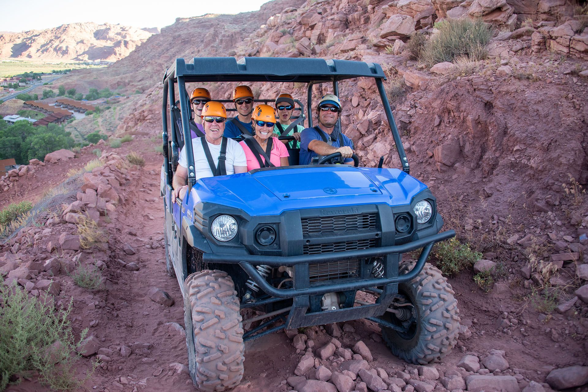 A blue utility vehicle with passengers wearing helmets travels on a dirt trail in a rocky canyon.