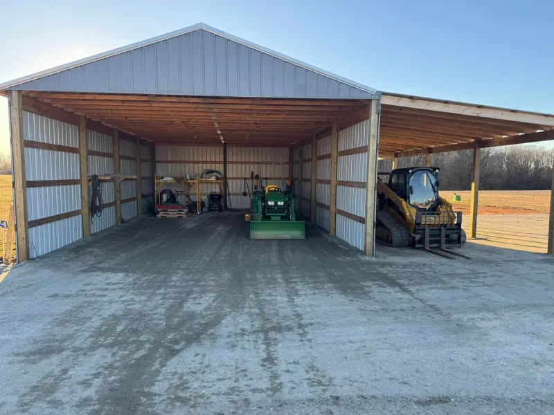 Open-sided farm shed with a green tractor and skid steer parked inside on a concrete floor