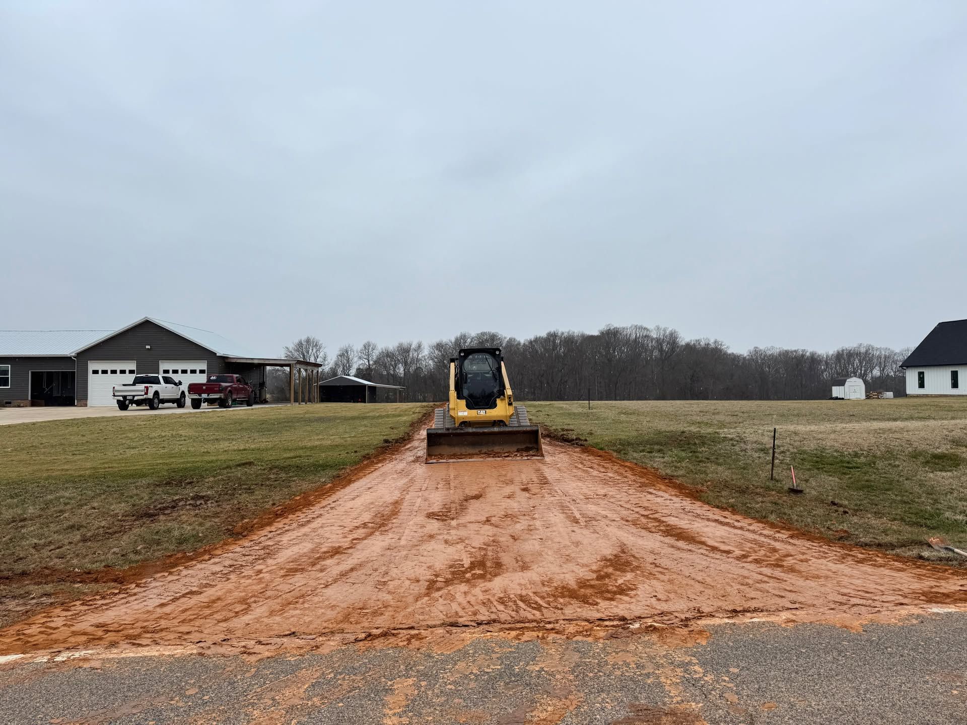 Yellow bulldozer grading a red dirt driveway between houses on a cloudy day