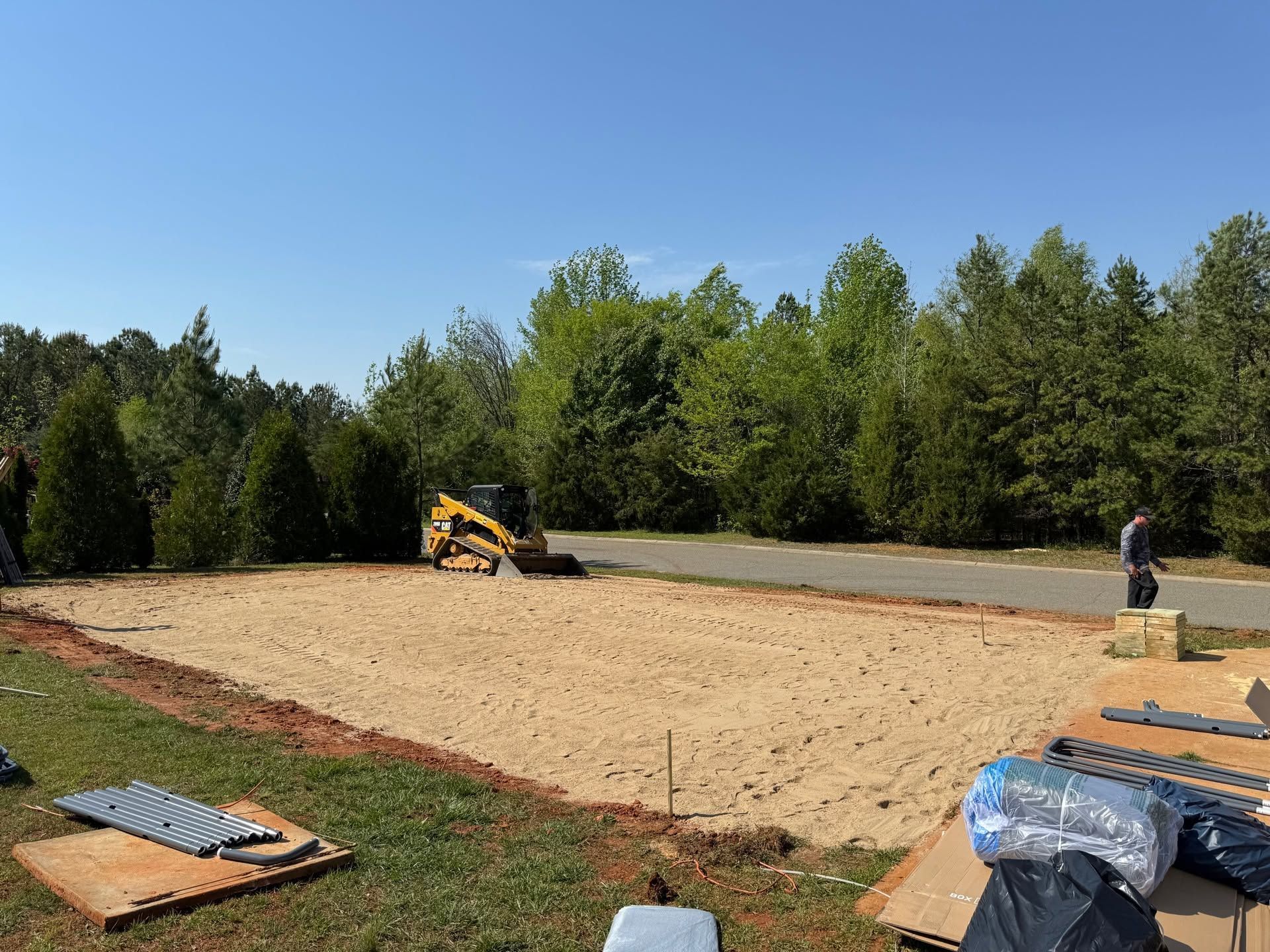 Outdoor worksite with a small bulldozer leveling a gravel pad beside trees and a person standing nearby.