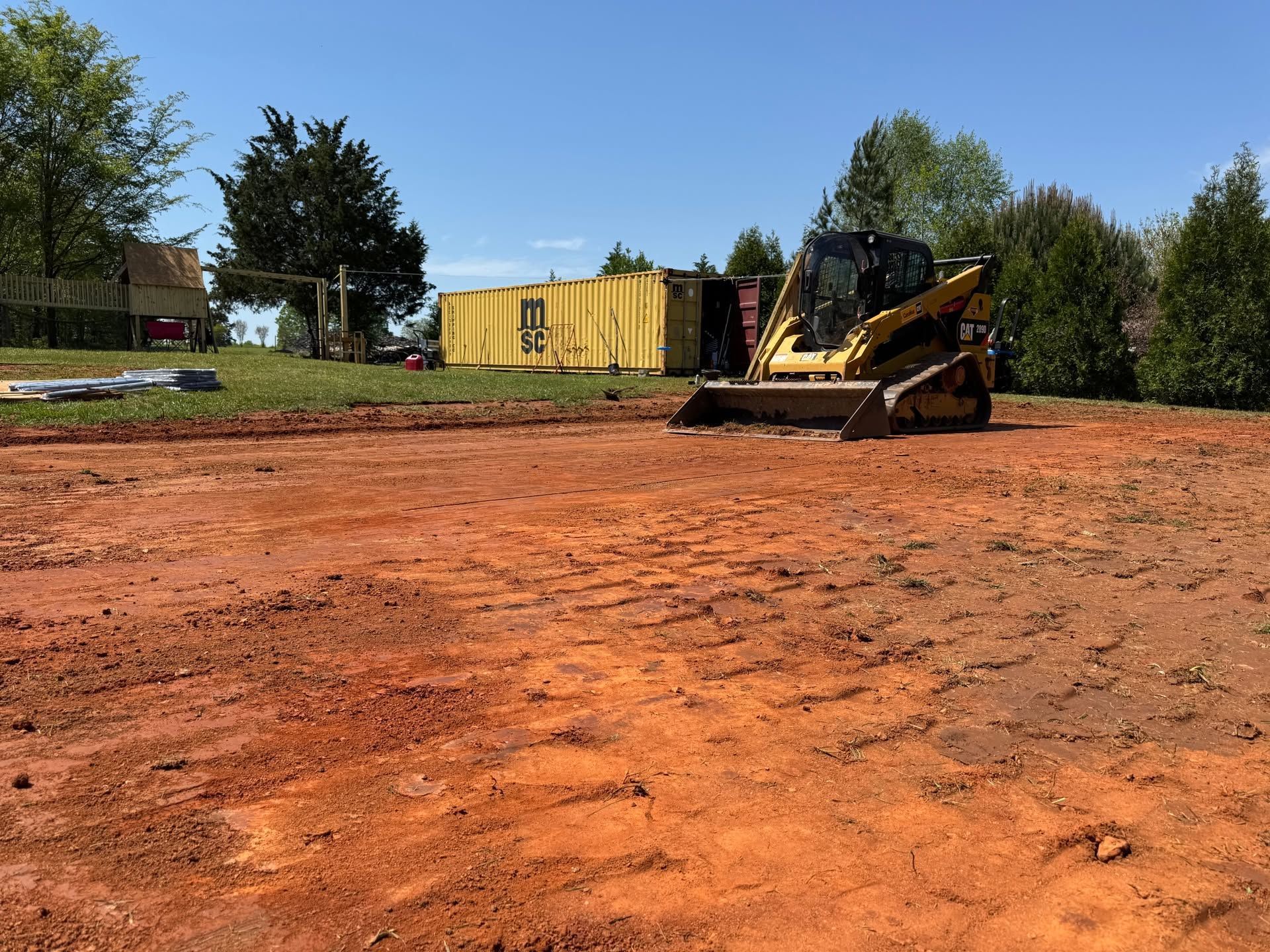 Bulldozer grading red dirt lot with a yellow container and trees in the background