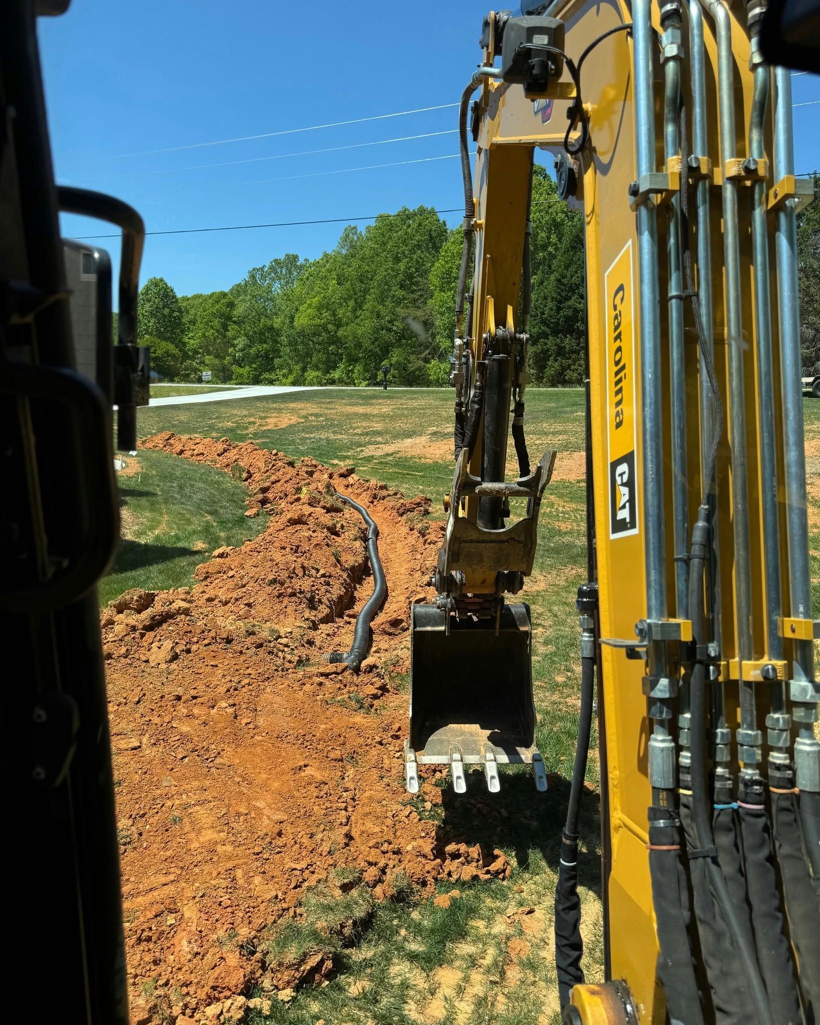 Yellow excavator digging a dirt path beside a grassy field under a clear blue sky