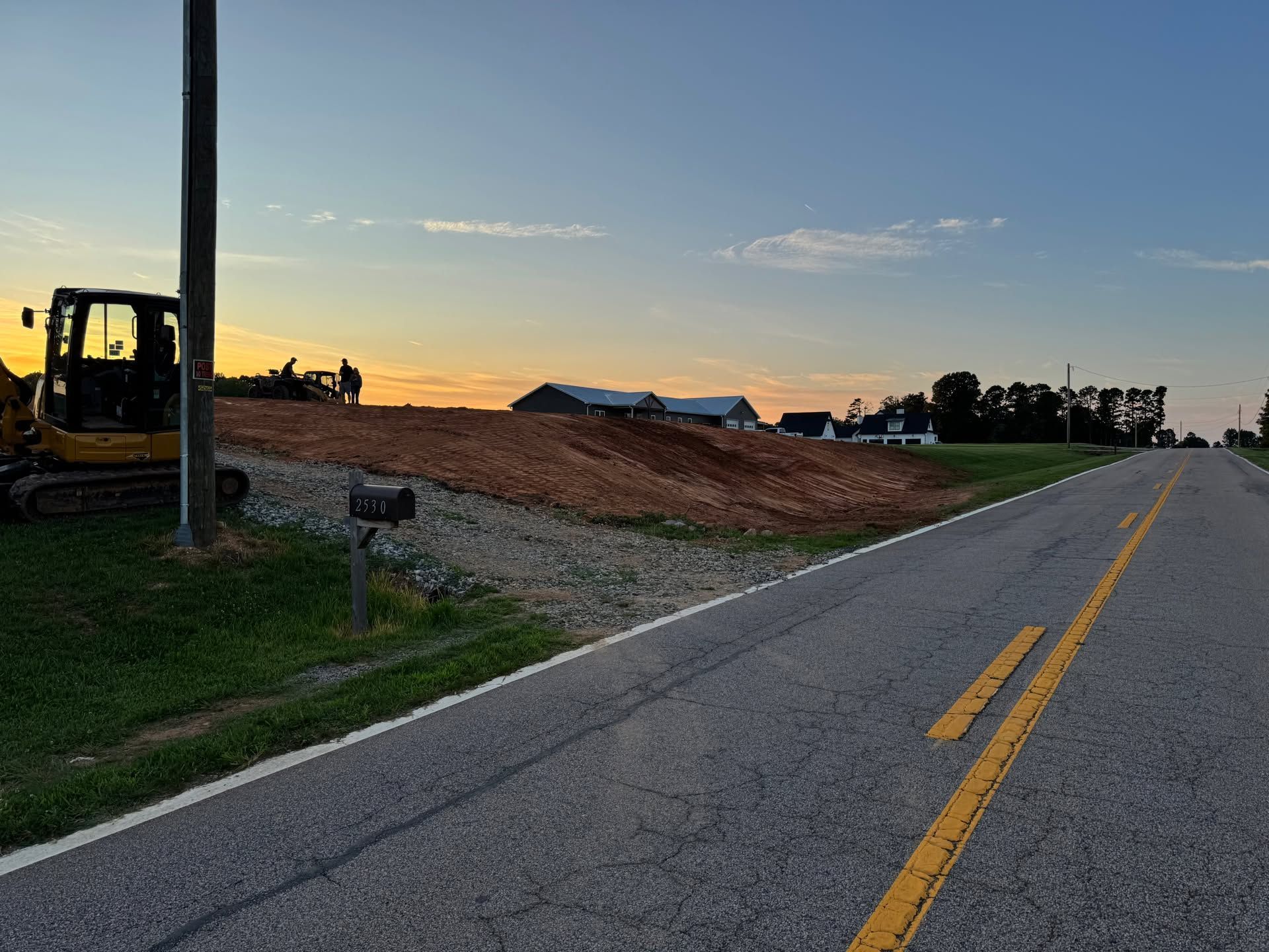 Roadside construction site at sunset with excavator, dirt mound, and empty two-lane road.