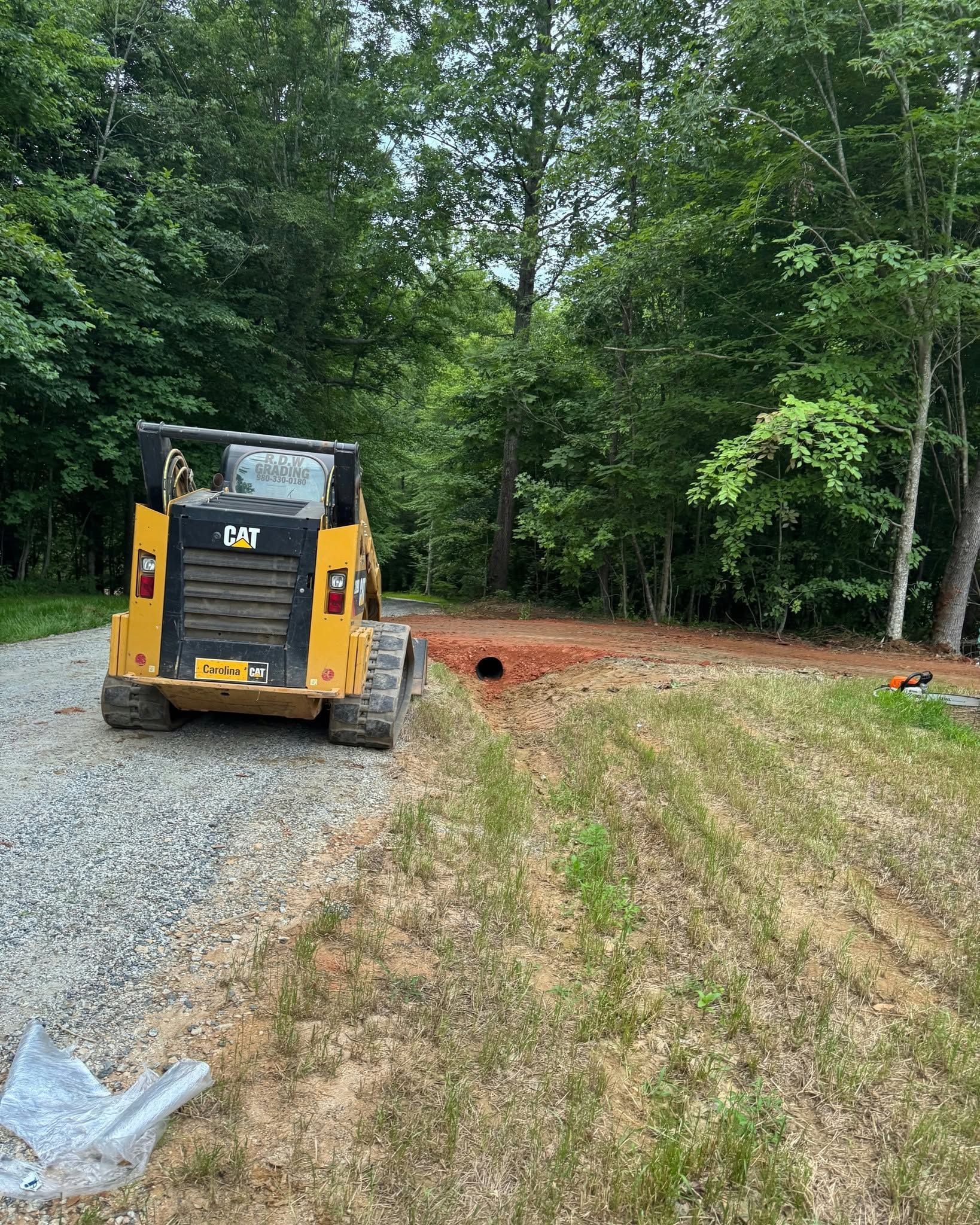 Yellow skid steer on gravel by a wooded clearing with a dirt path and drainage pipe