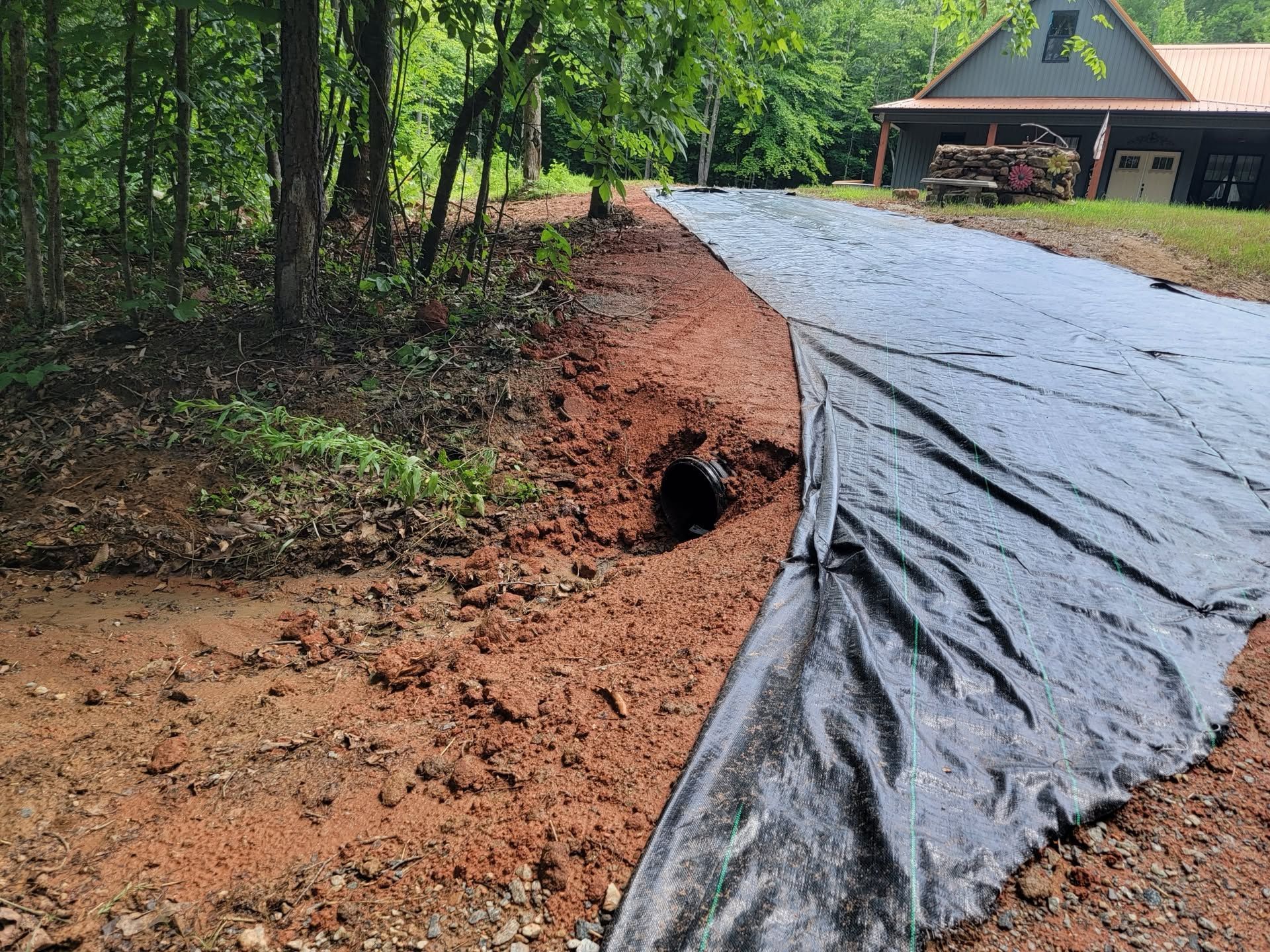 Black tarp lining a muddy roadside erosion trench beside a wooded driveway and house