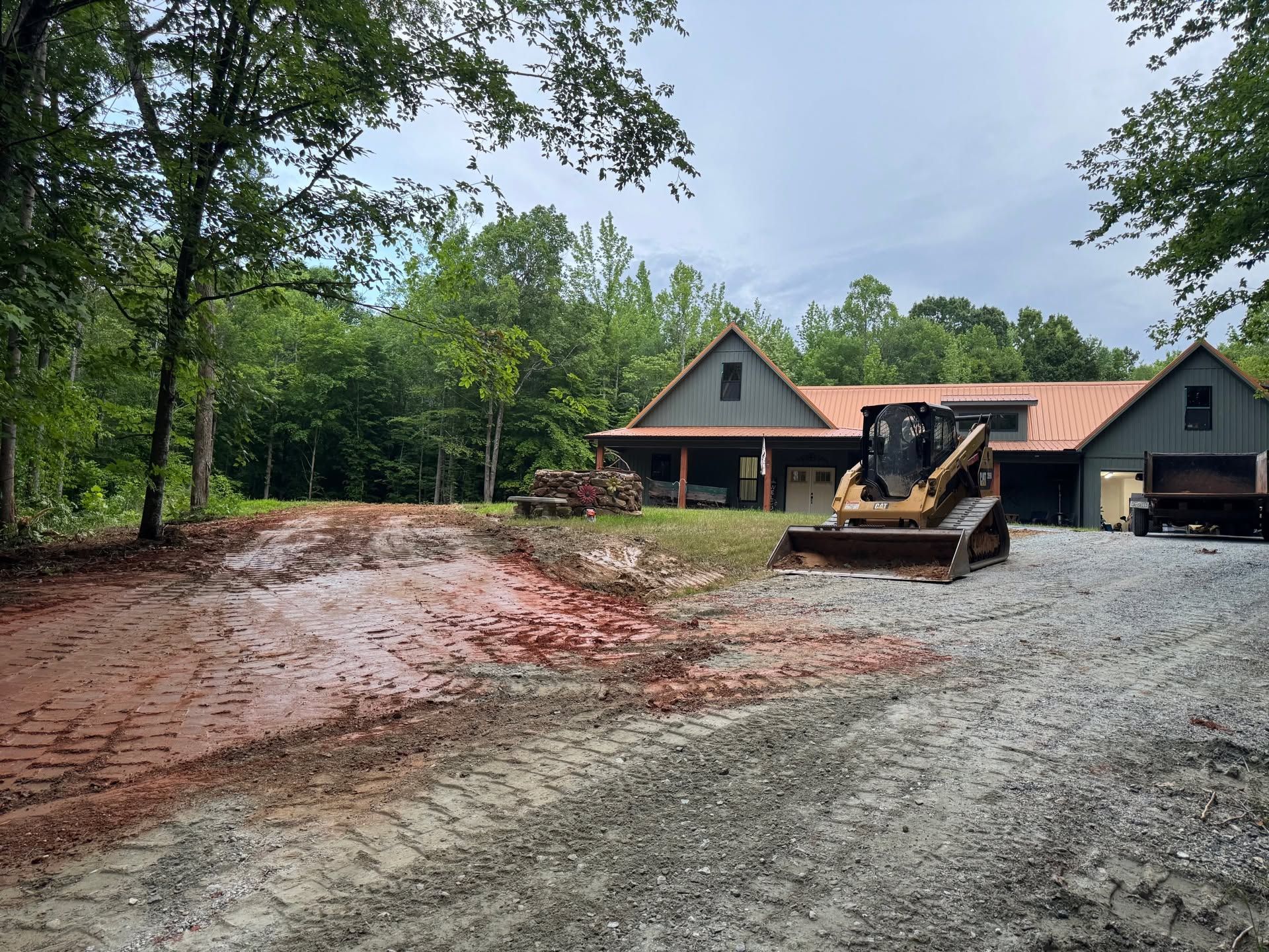 House under construction with a bulldozer on a muddy gravel driveway surrounded by trees