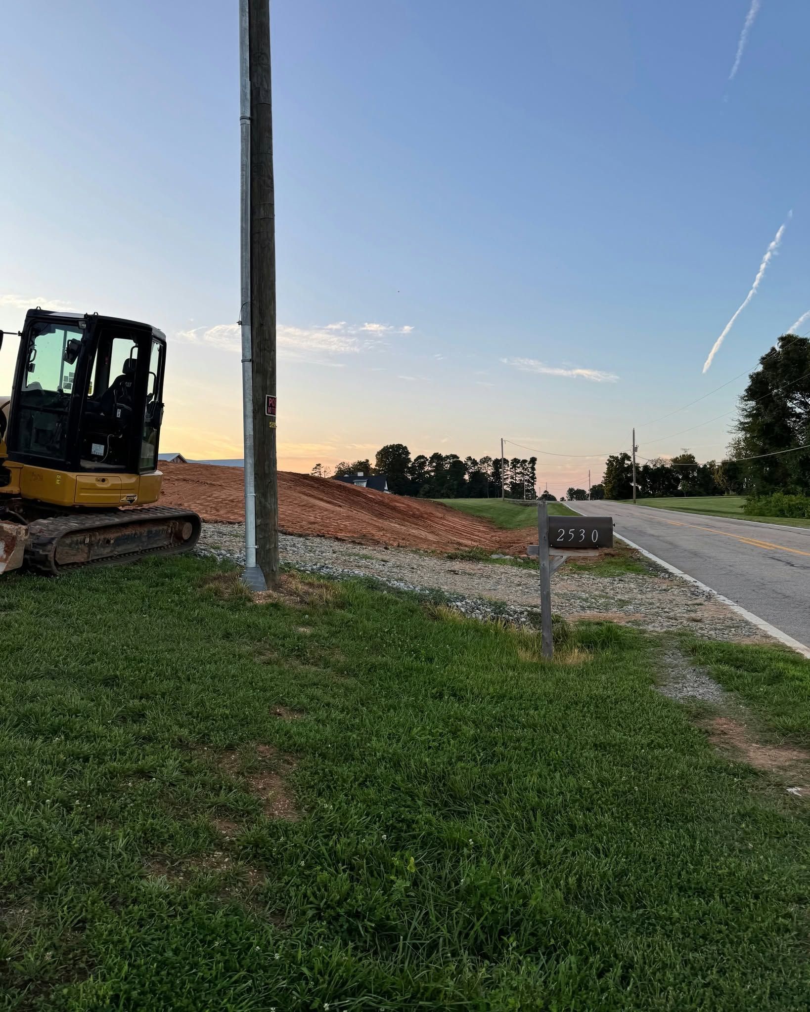 Construction site beside road at dusk, with excavator, dirt pile, utility pole, and grassy foreground.