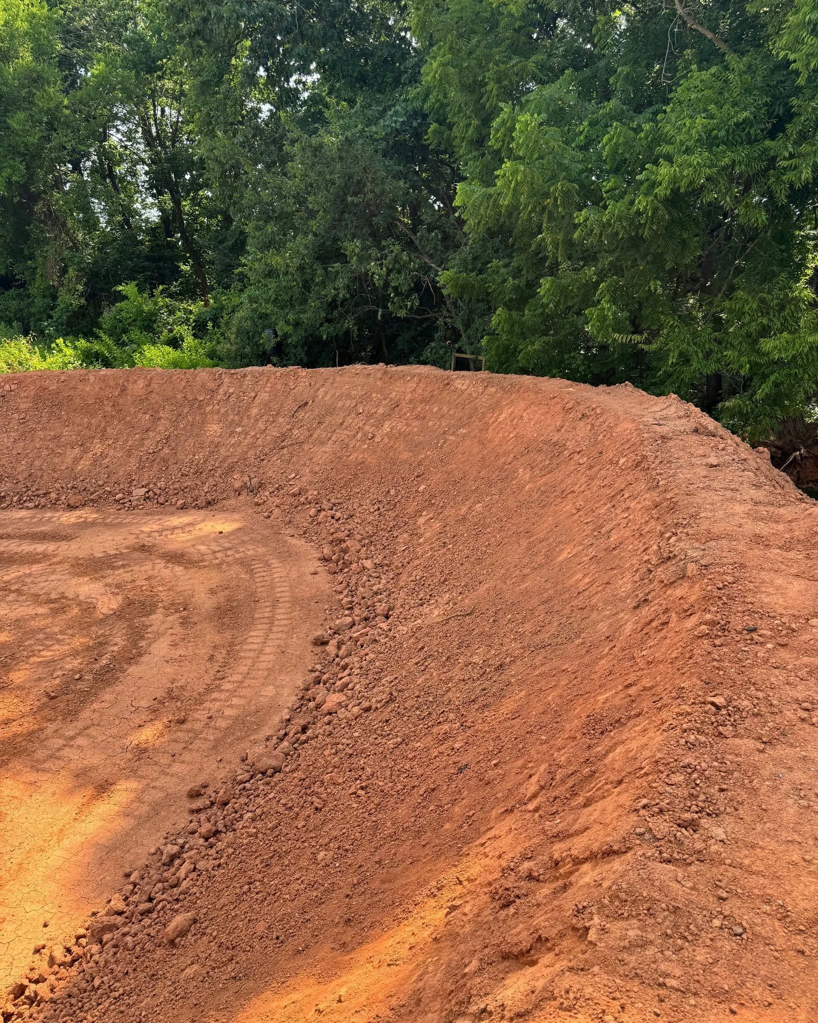 A large reddish dirt mound beside dense green trees outdoors