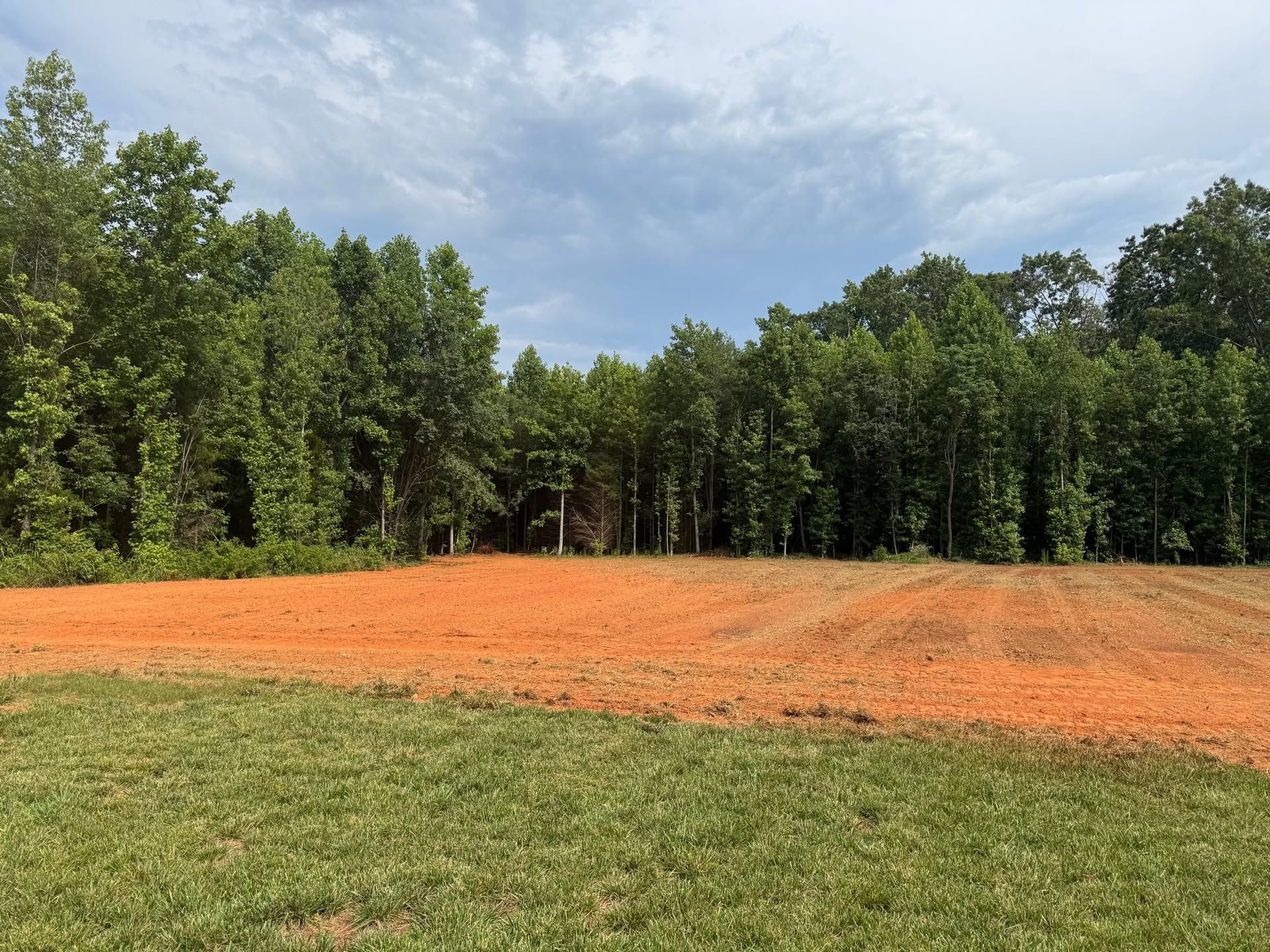 Grassy field with a large orange dirt clearing bordered by dense green trees under a partly cloudy sky