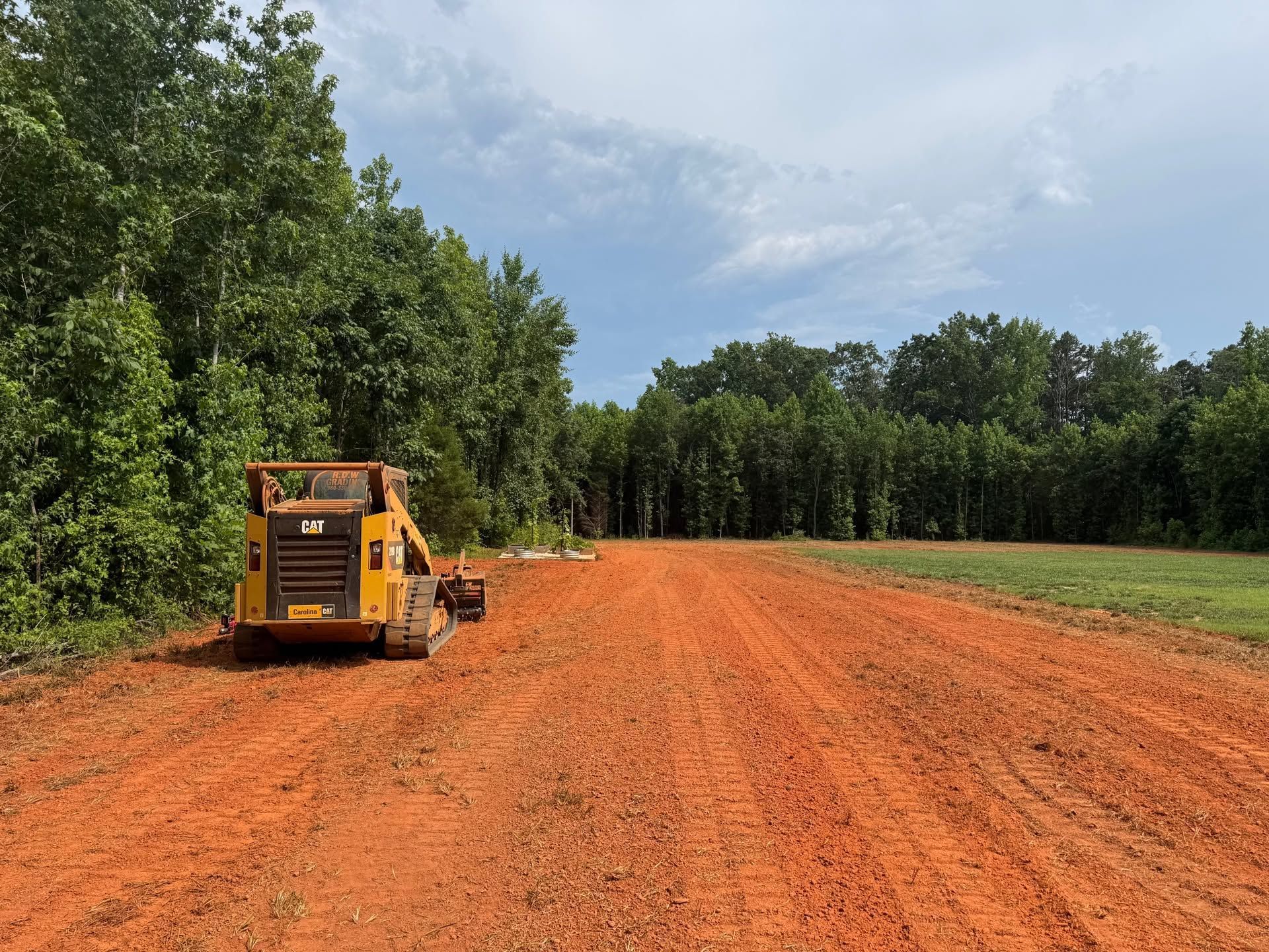 Yellow road grader on a red dirt road beside green trees under a cloudy sky