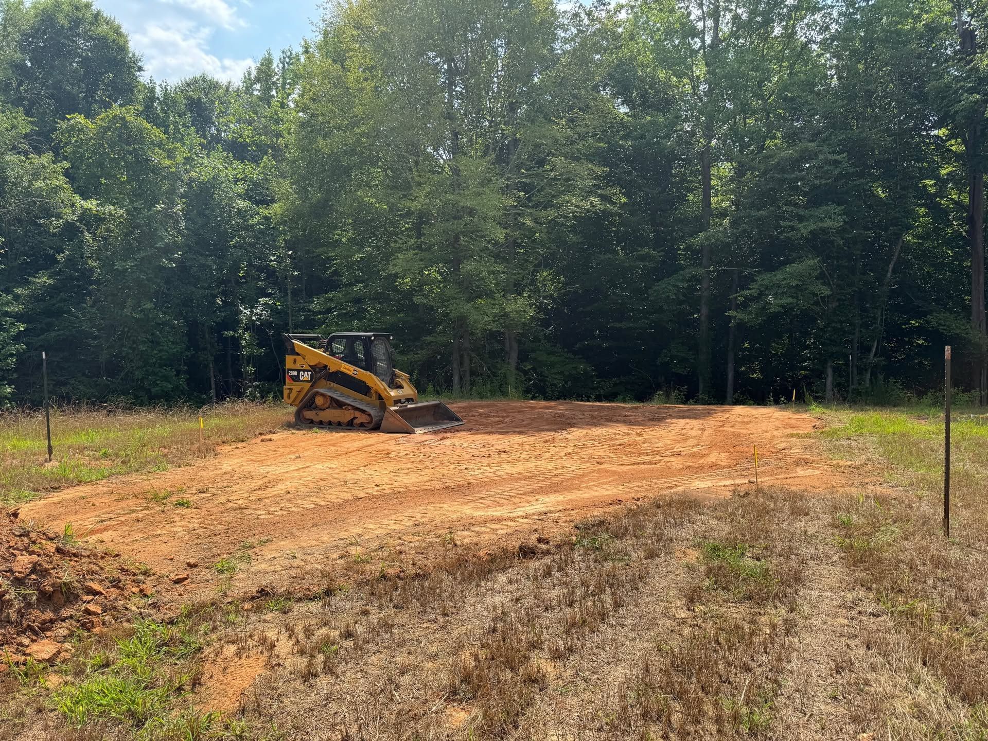 Yellow skid steer clearing a dirt lot beside trees and a fenced wooded area