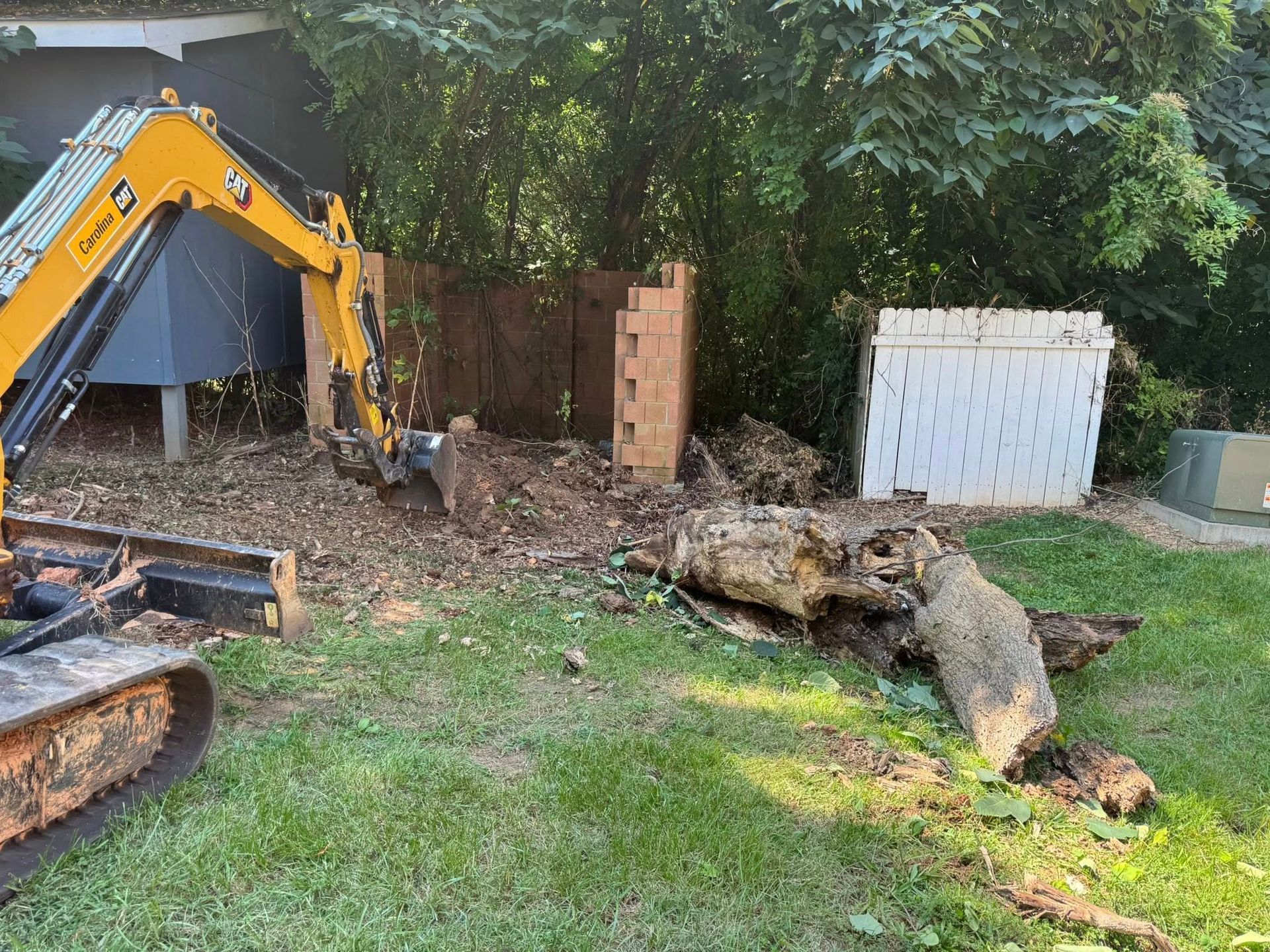 Yellow excavator removing a large tree stump in a backyard beside a wooden fence and shed.