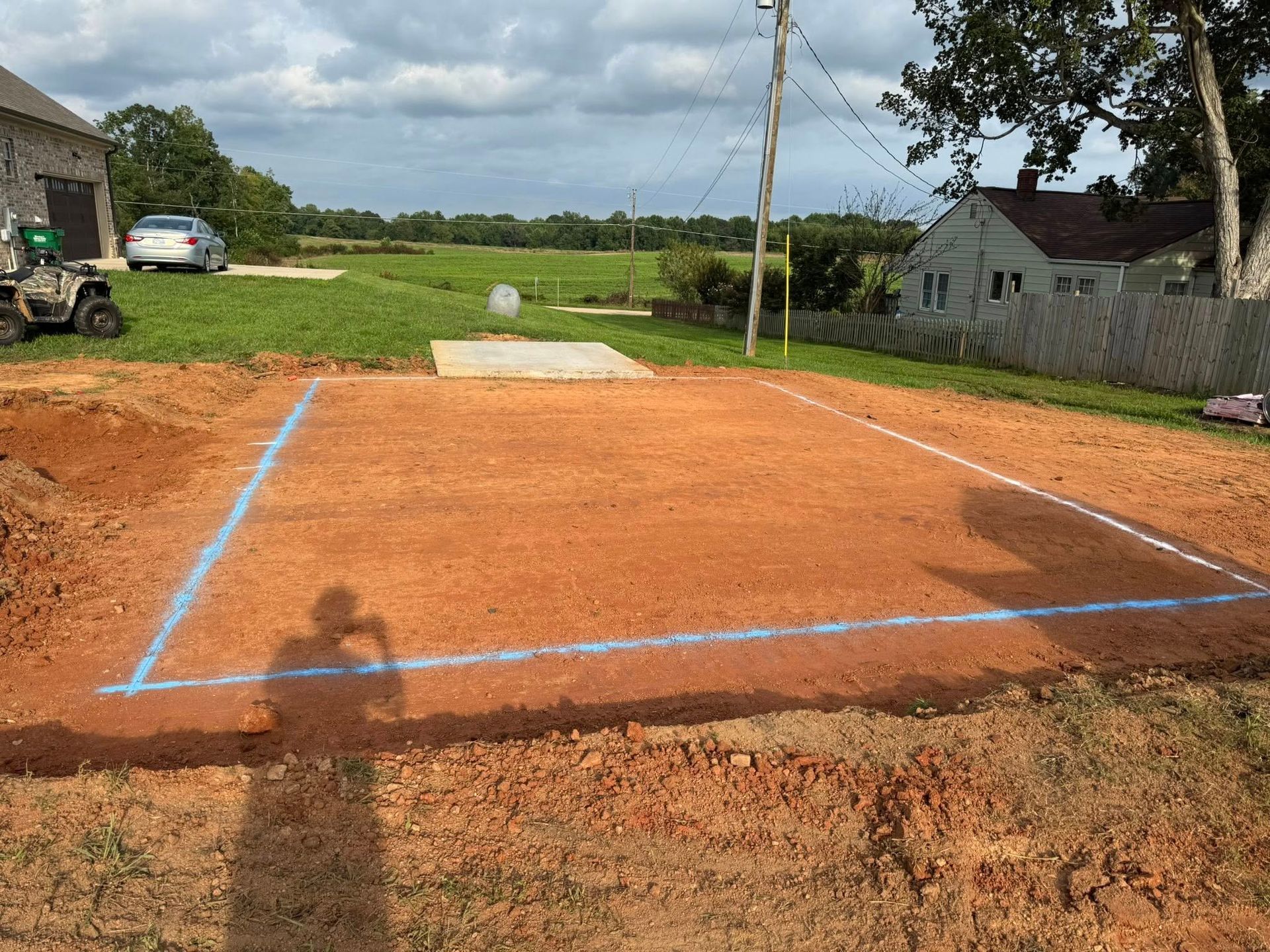 Blue-outlined dirt foundation pad at a rural construction site under cloudy skies