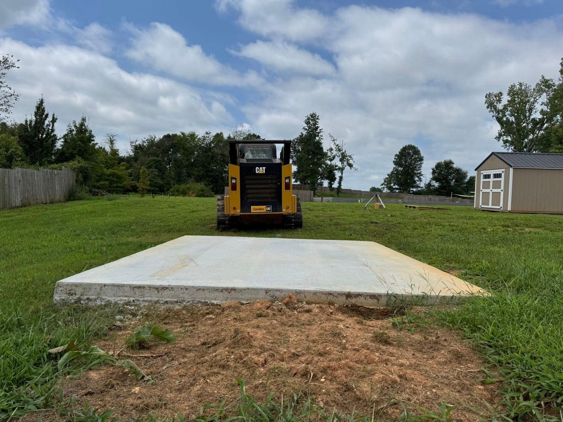 Yellow construction machine parked on a concrete slab in a grassy yard under a cloudy sky