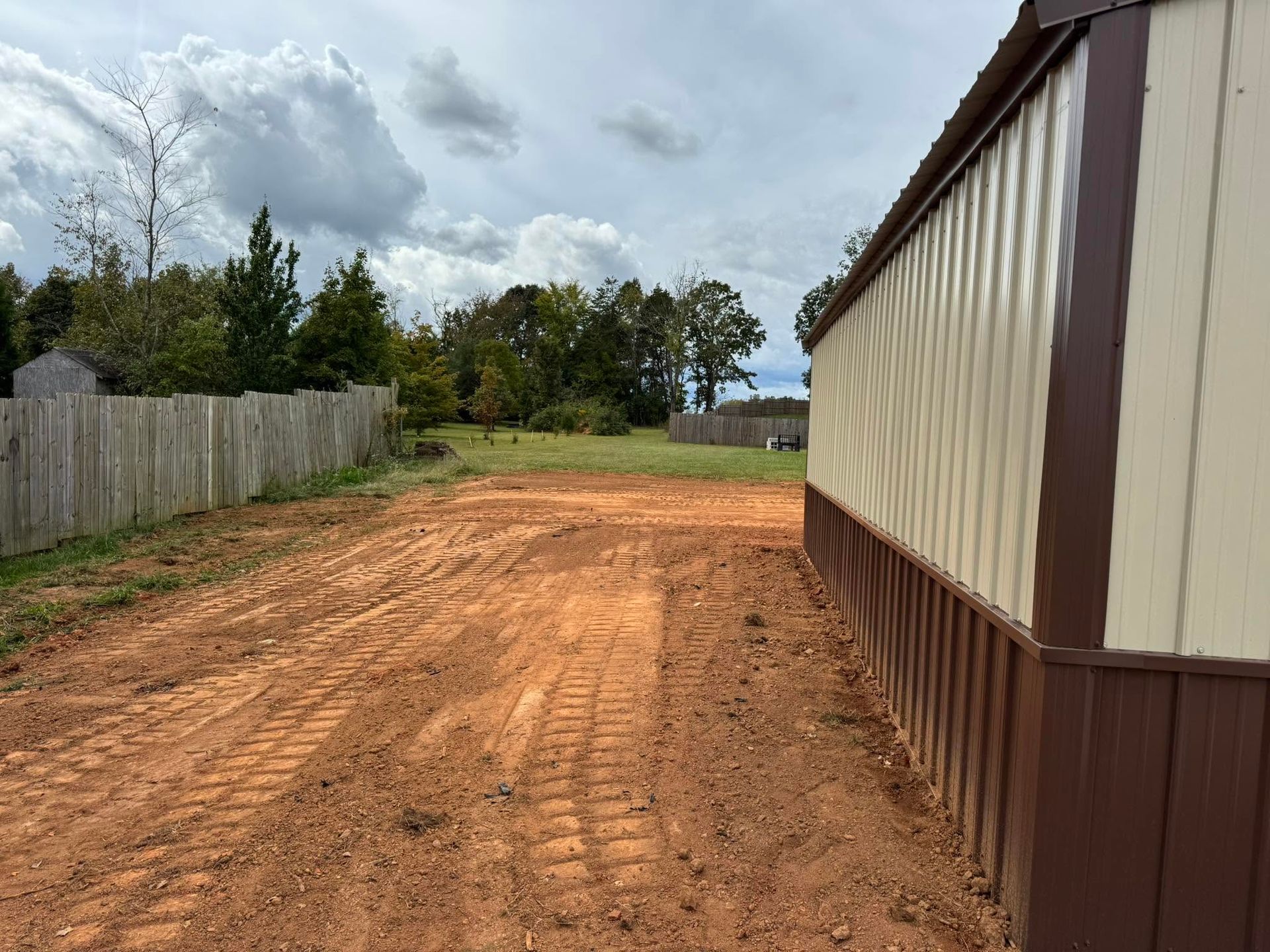 Dirt path beside a beige metal building, with a fenced yard and cloudy sky beyond.