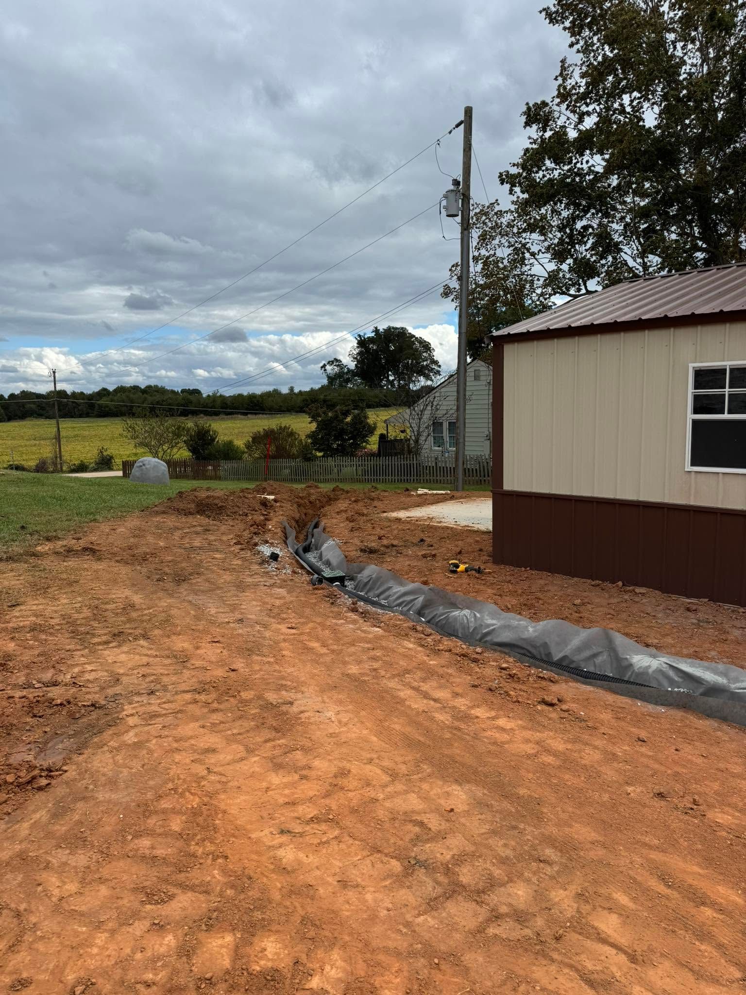 Dirt road beside a beige building, with utility pole, black tarp edging, and cloudy rural landscape.