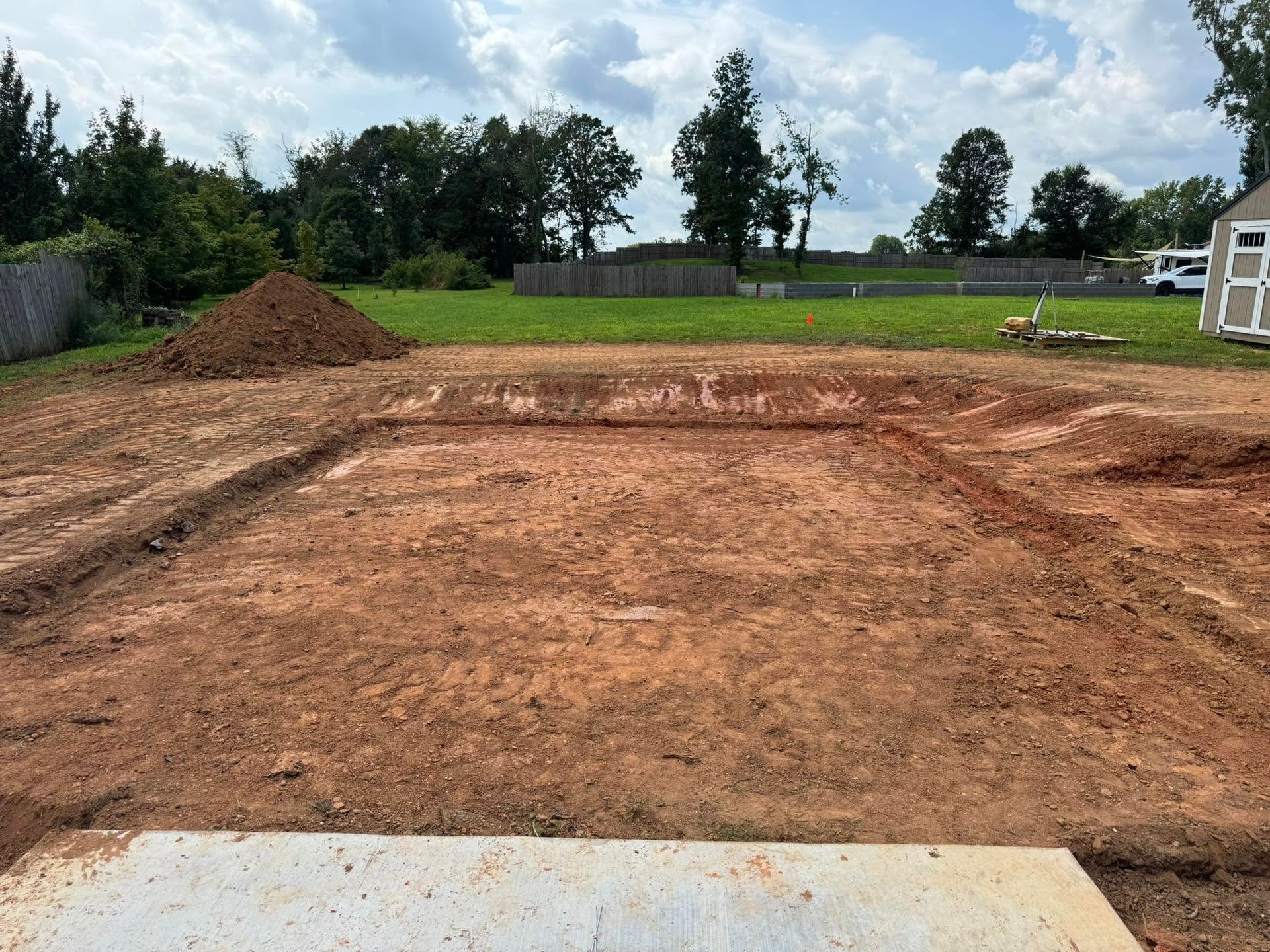 Cleared dirt lot with graded soil, a mound of earth, and trees under a cloudy sky