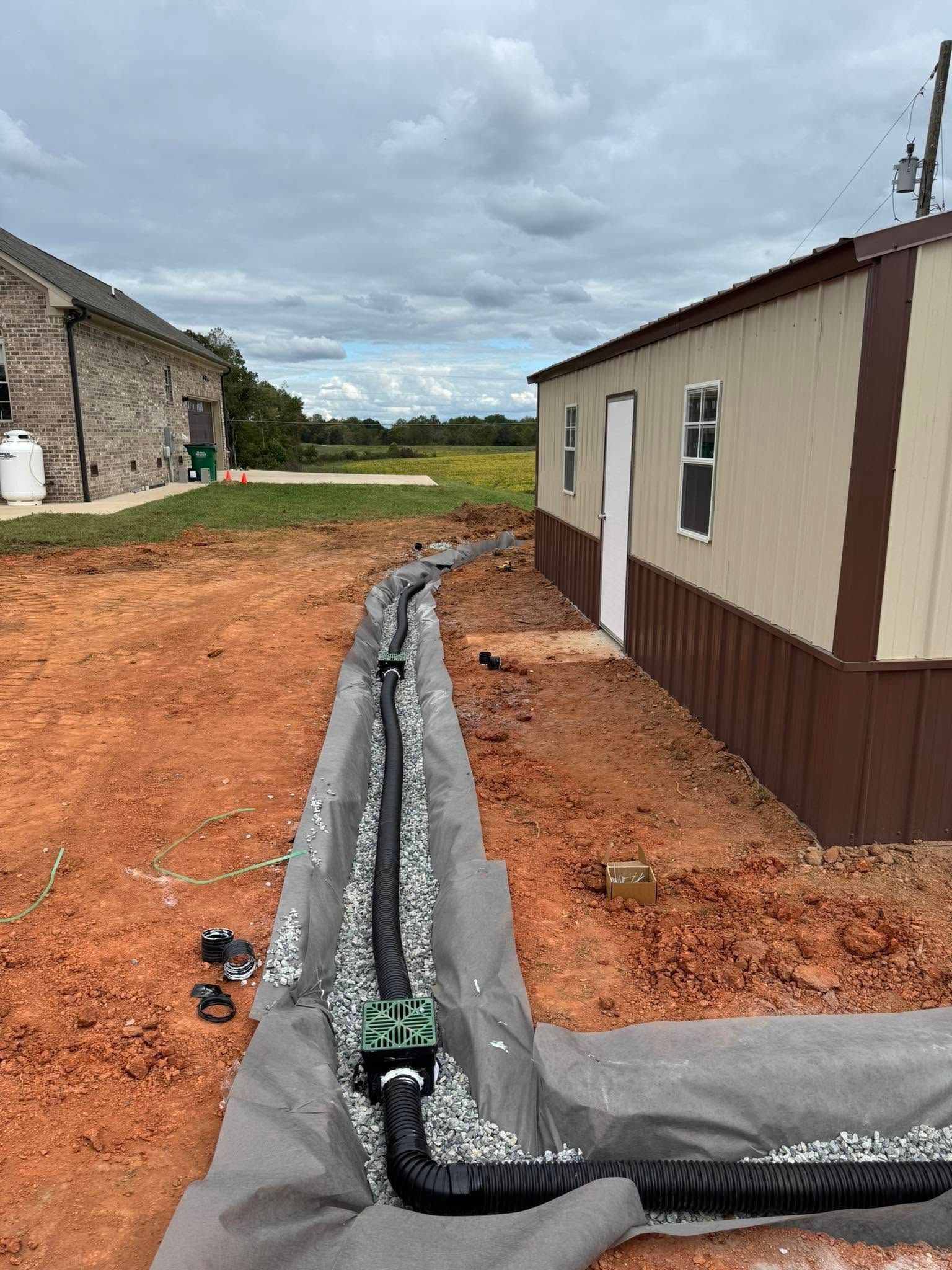 Gravel trench with black drainage pipe between two mobile homes on a red dirt lot