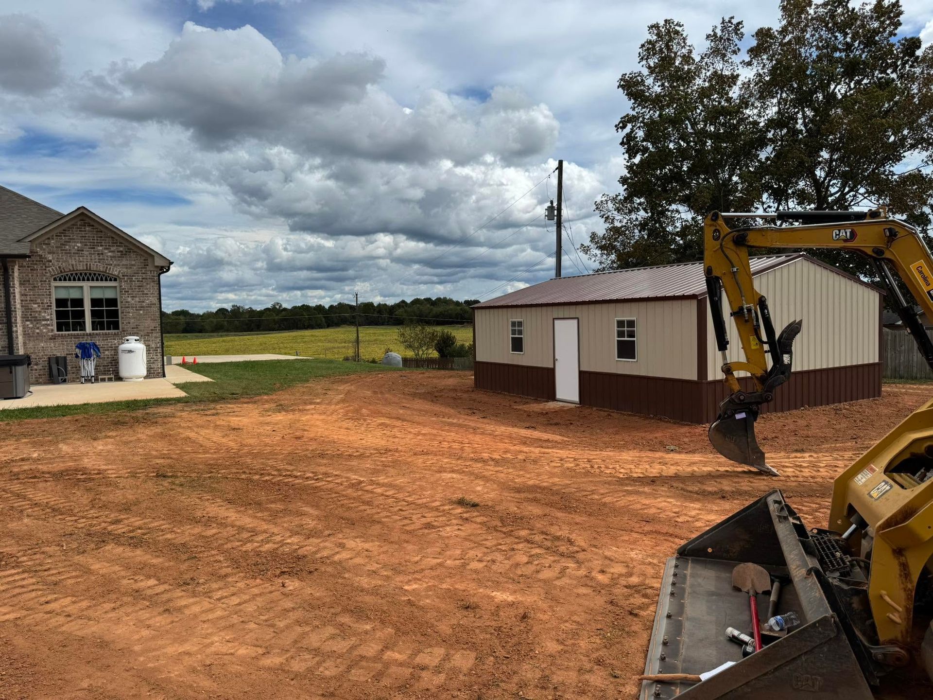 Construction site with excavator beside a small building and a larger stone house under cloudy skies