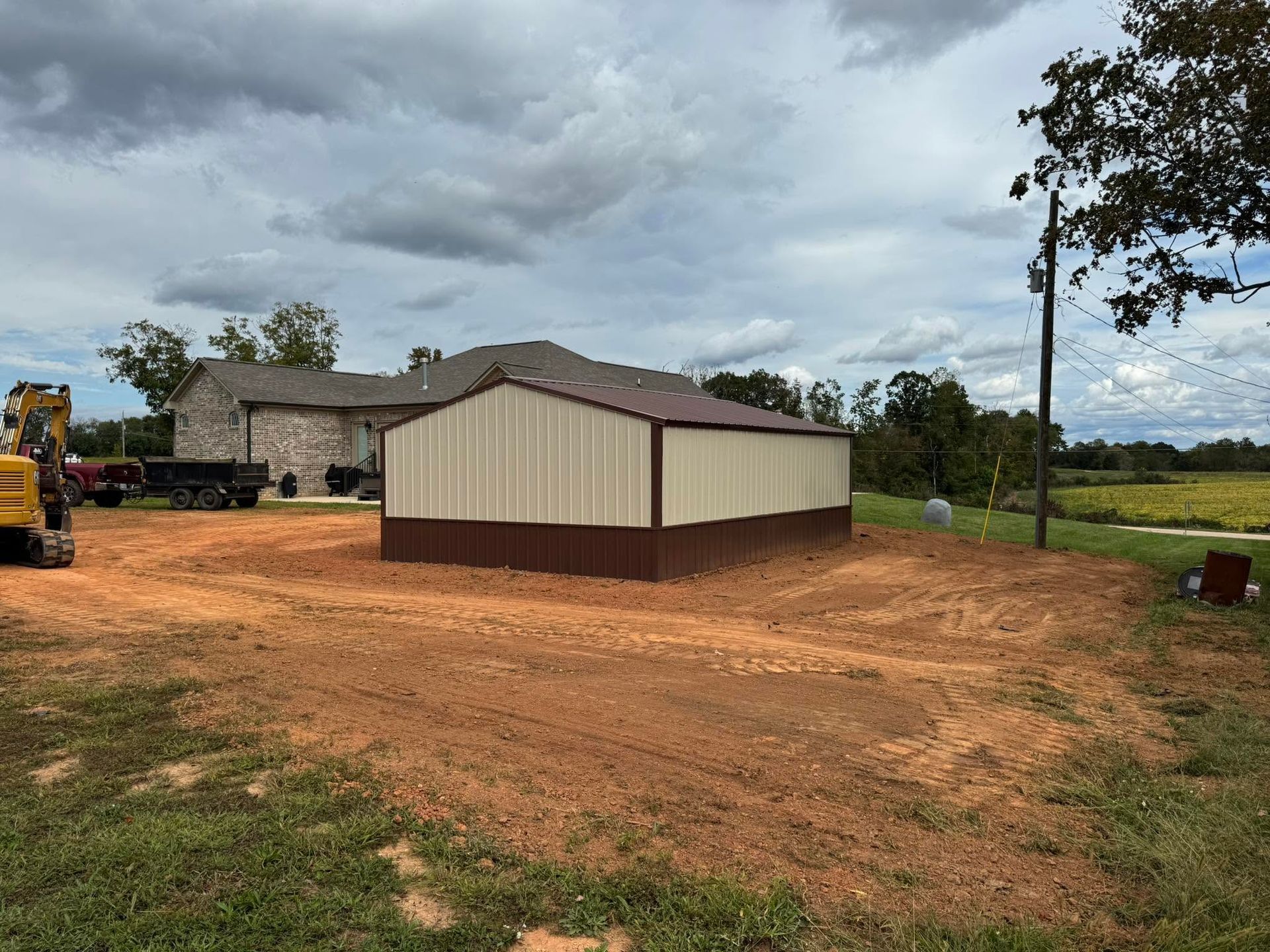 Small beige-and-brown shed on a cleared dirt lot under cloudy skies, with a house and construction equipment nearby