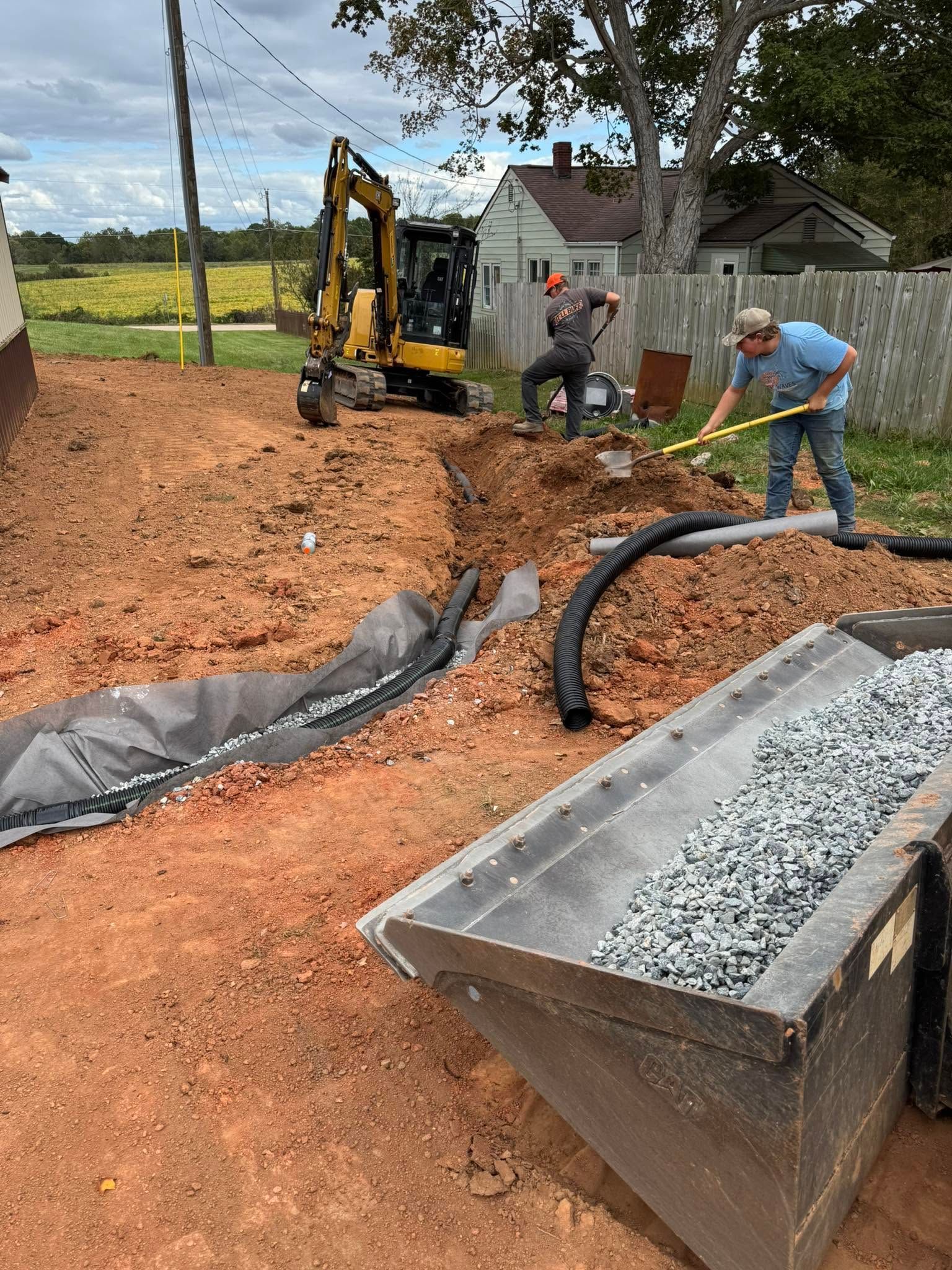 Workers grading a construction site beside a loader and gravel bin under cloudy skies