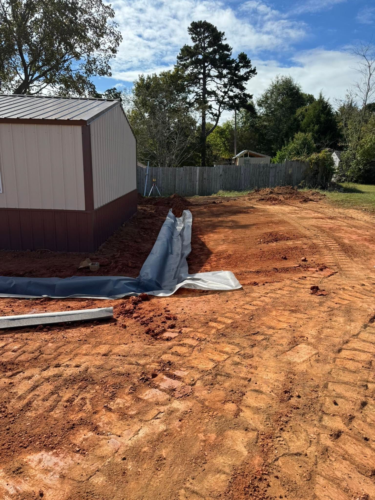 Dirt lot beside a shed, with red clay ground, a tarp-covered edge, and trees and fence in the background