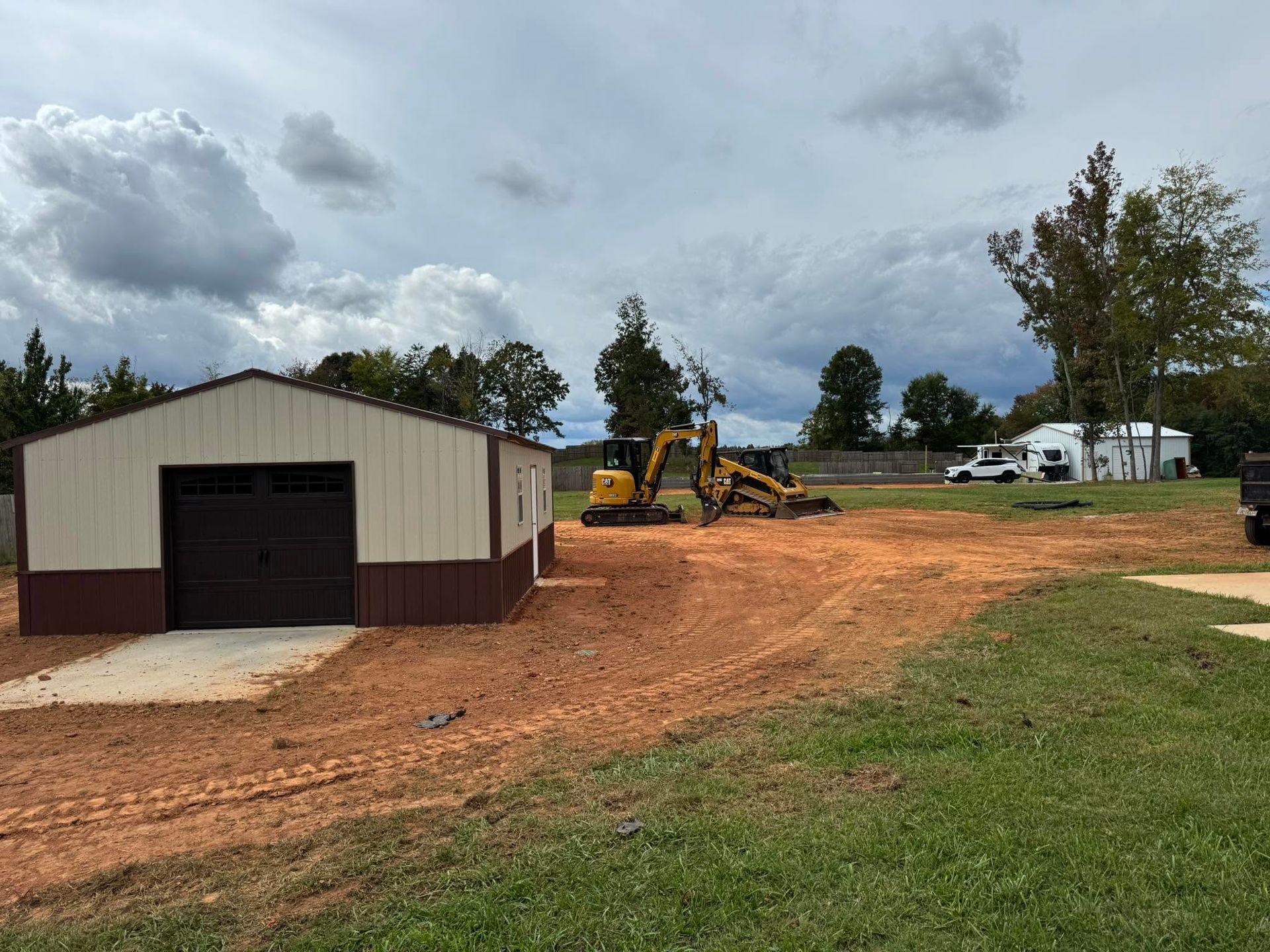 Rural worksite with a metal shed, excavator, dirt driveway, and white truck under a cloudy sky