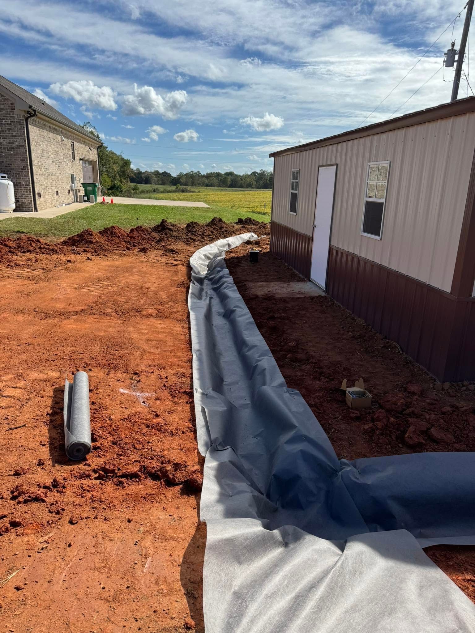 Gravel trench with black liner beside a beige building, under construction in a sunny rural area