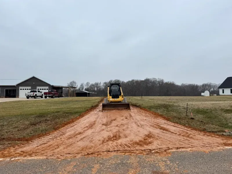 Yellow skid steer on a dirt driveway under construction in a grassy field near houses