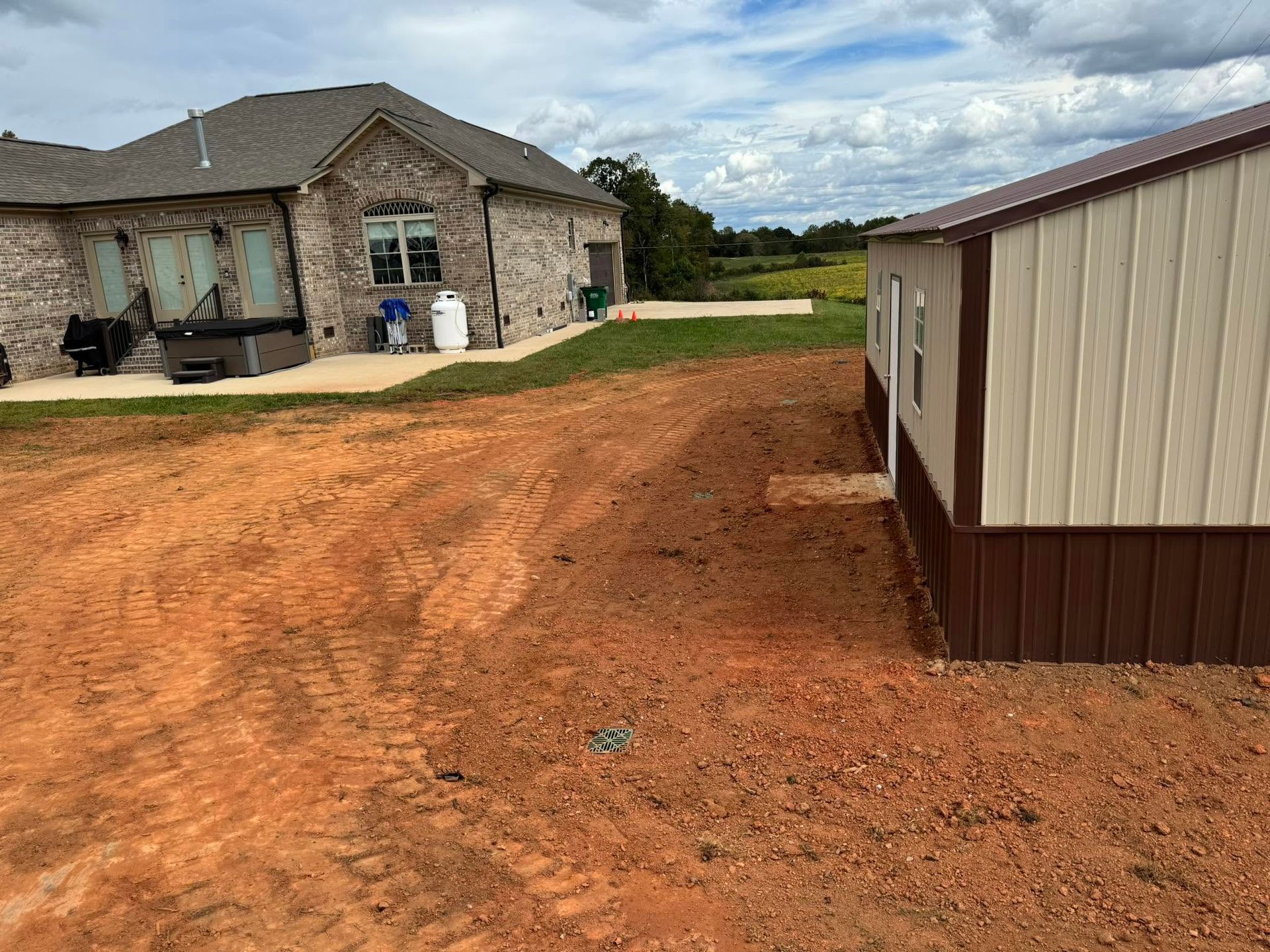 Rural house with red dirt driveway and a metal outbuilding under a cloudy sky.