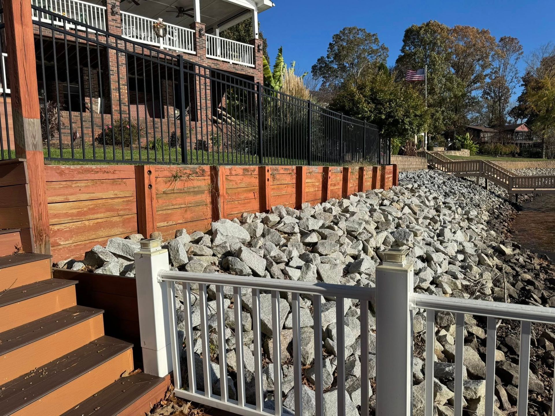 Riverside retaining wall under construction with gray rocks, brick steps, and a black metal fence by a house