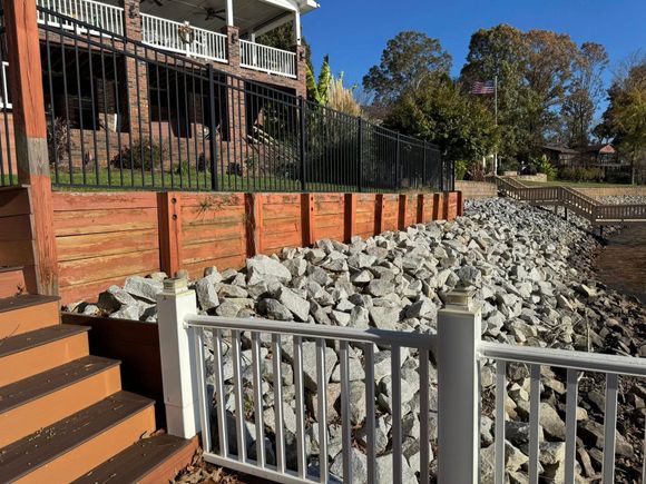 Rock-lined drainage channel beside a brick retaining wall and house fence on a sunny day