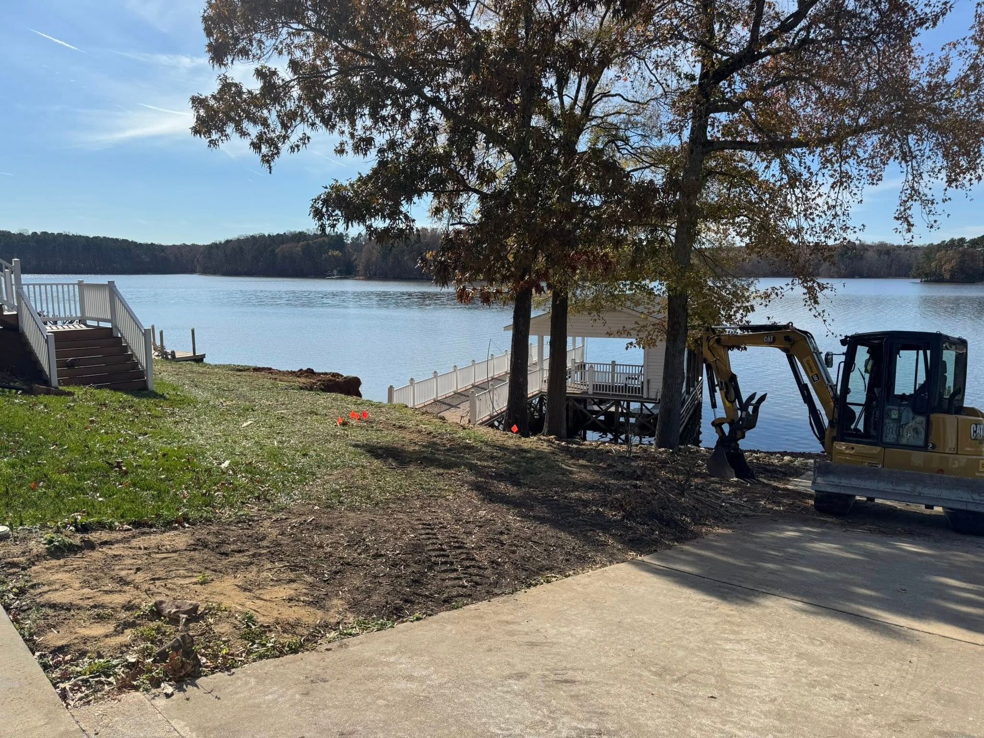 Lakeside shoreline with dock, trees, and an excavator beside a concrete path